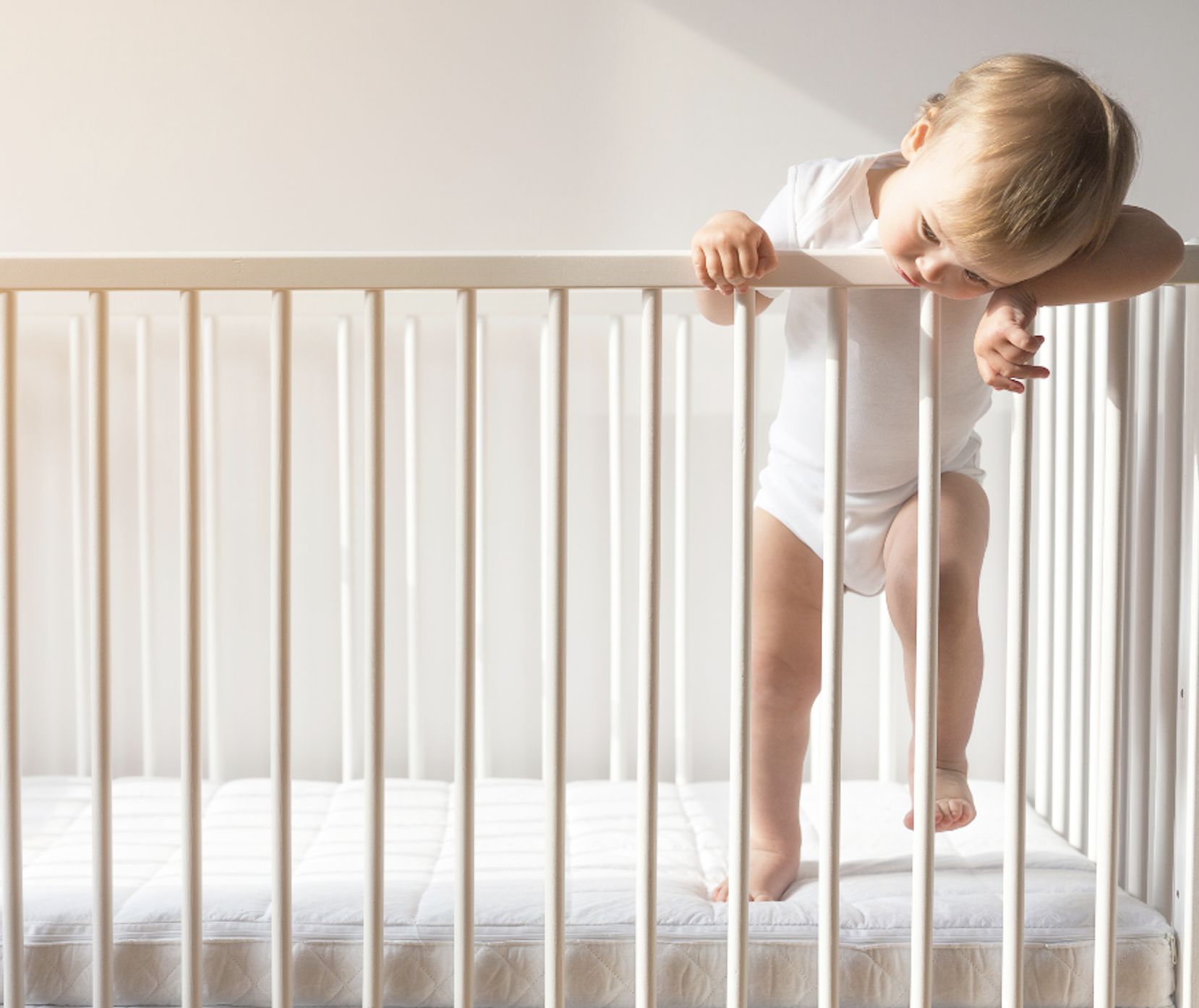 A baby stands alone in a crib in a lit room, head resting on the crib rail, looking tired but unable to settle. A familiar scene for families struggling with sleep training that isn't working.
