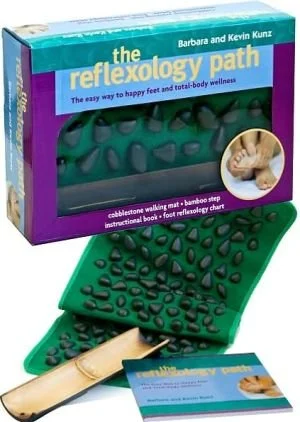 The image shows a boxed set of a reflexology path kit, including a cobblestone walking mat, a bamboo step, an instructional book, and a foot reflexology chart. The packaging is purple and green, with the title 'the reflexology path' and includes promotional text emphasizing foot and body wellness.