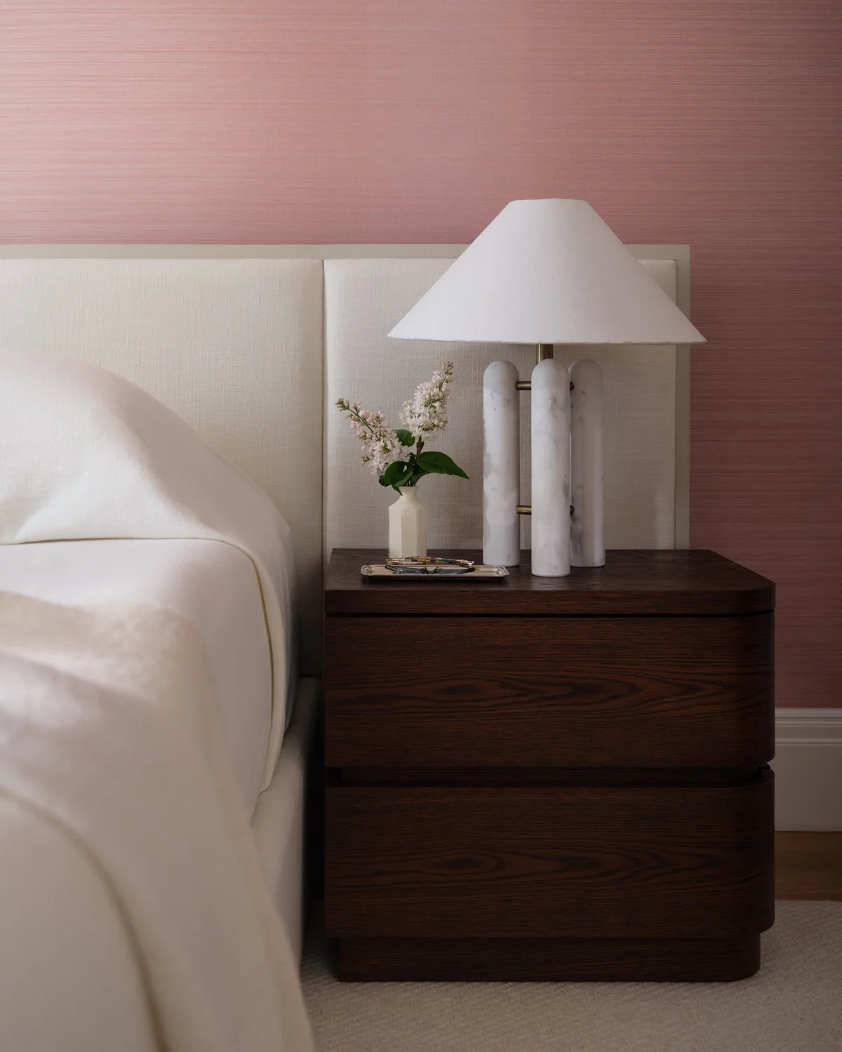 Nightstand with a lamp, a small vase with flowers, and glasses in front of a pink wall