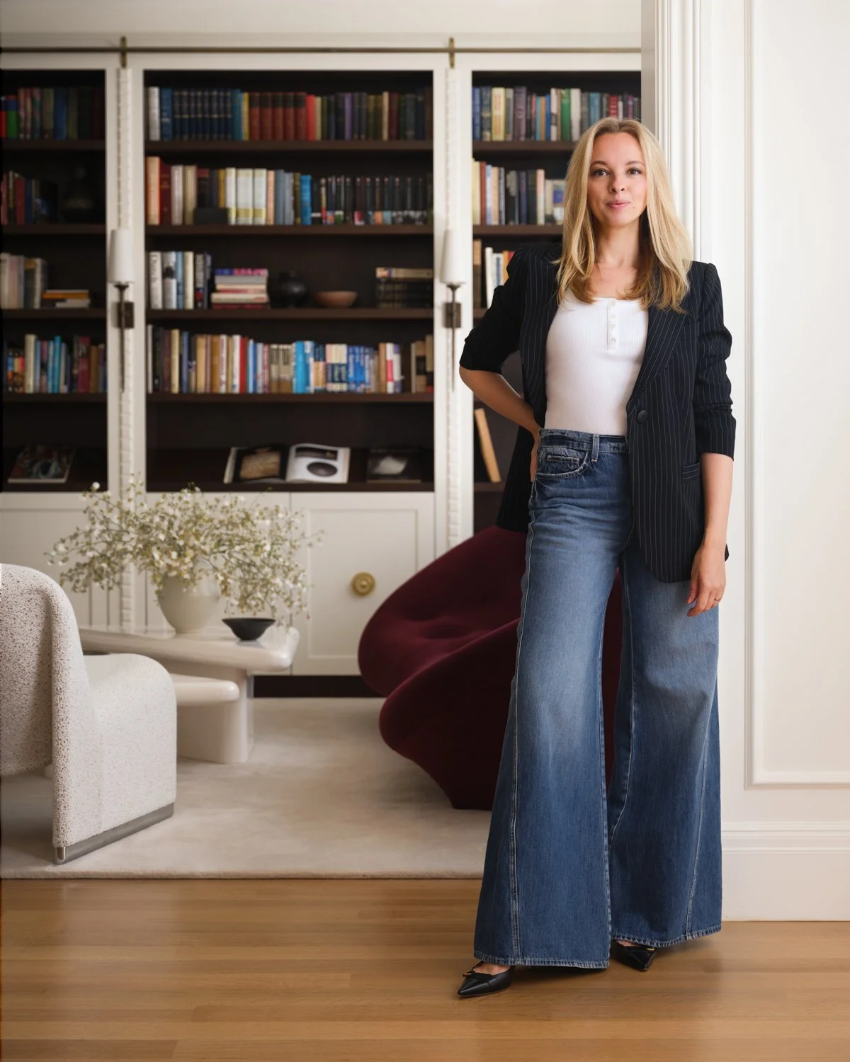A woman standing in a living room near a bookshelf, wearing a black blazer, white top, and wide-leg jeans.