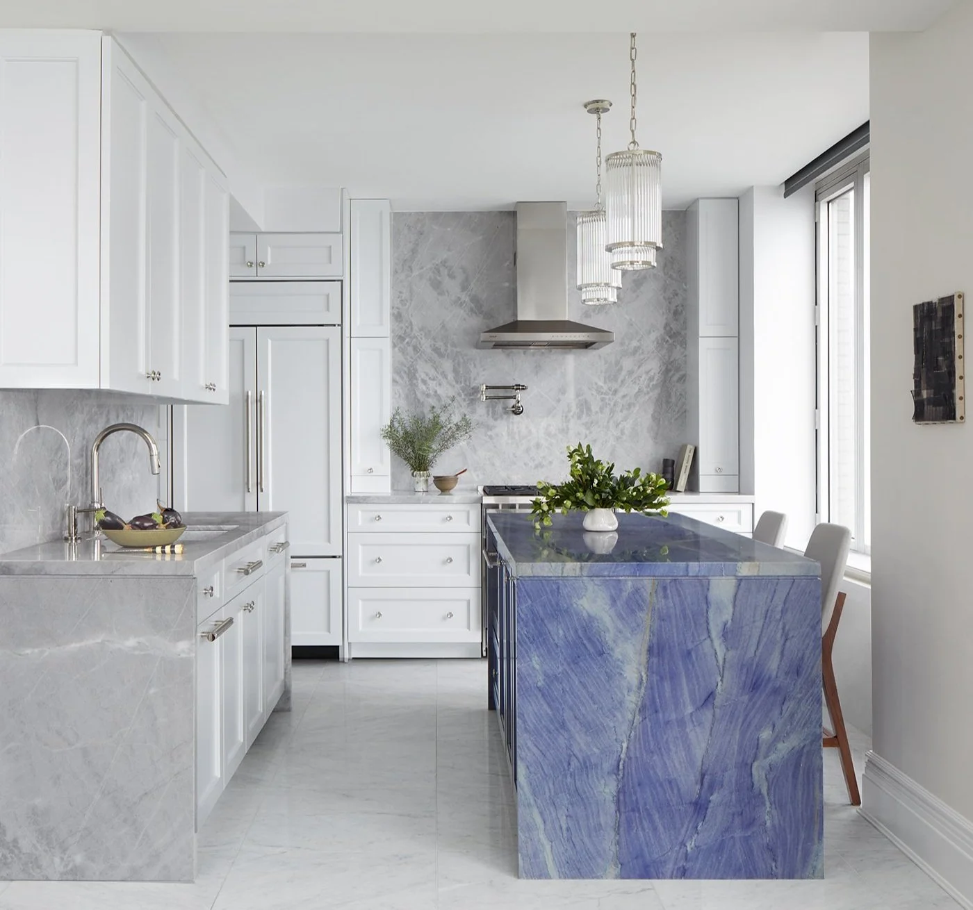 Modern kitchen with white cabinets, grey marble countertops, and a blue marble kitchen island. Hanging pendant lights, a window, and decorative green plants are also visible.