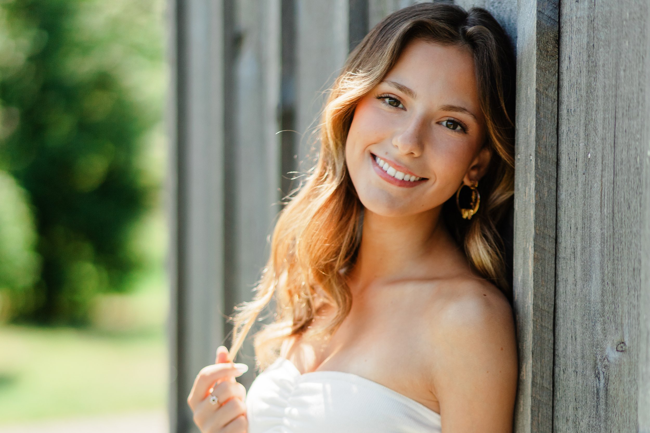 A young woman with long wavy hair, smiling, leaning against a wooden wall outdoors, wearing a white strapless top and hoop earrings.
