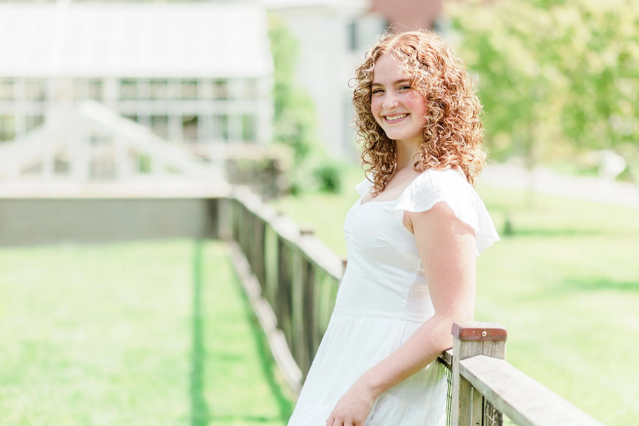 A young woman with curly red hair wearing a white dress, smiling and standing outdoors next to a wooden fence with a blurred background of greenery and buildings.