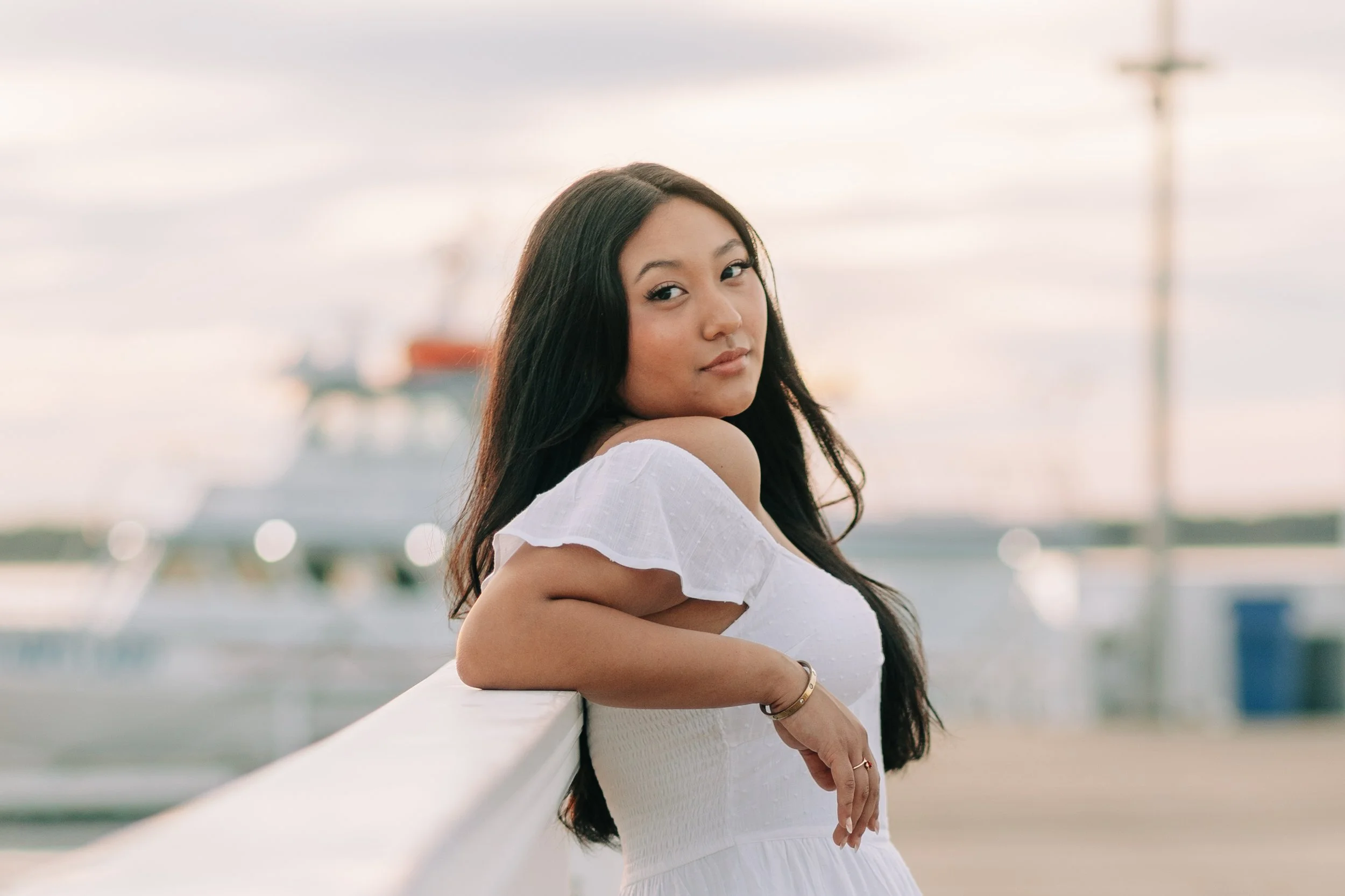 Young woman with long dark hair leaning on a white railing at the waterfront during sunset, with boats in the background.