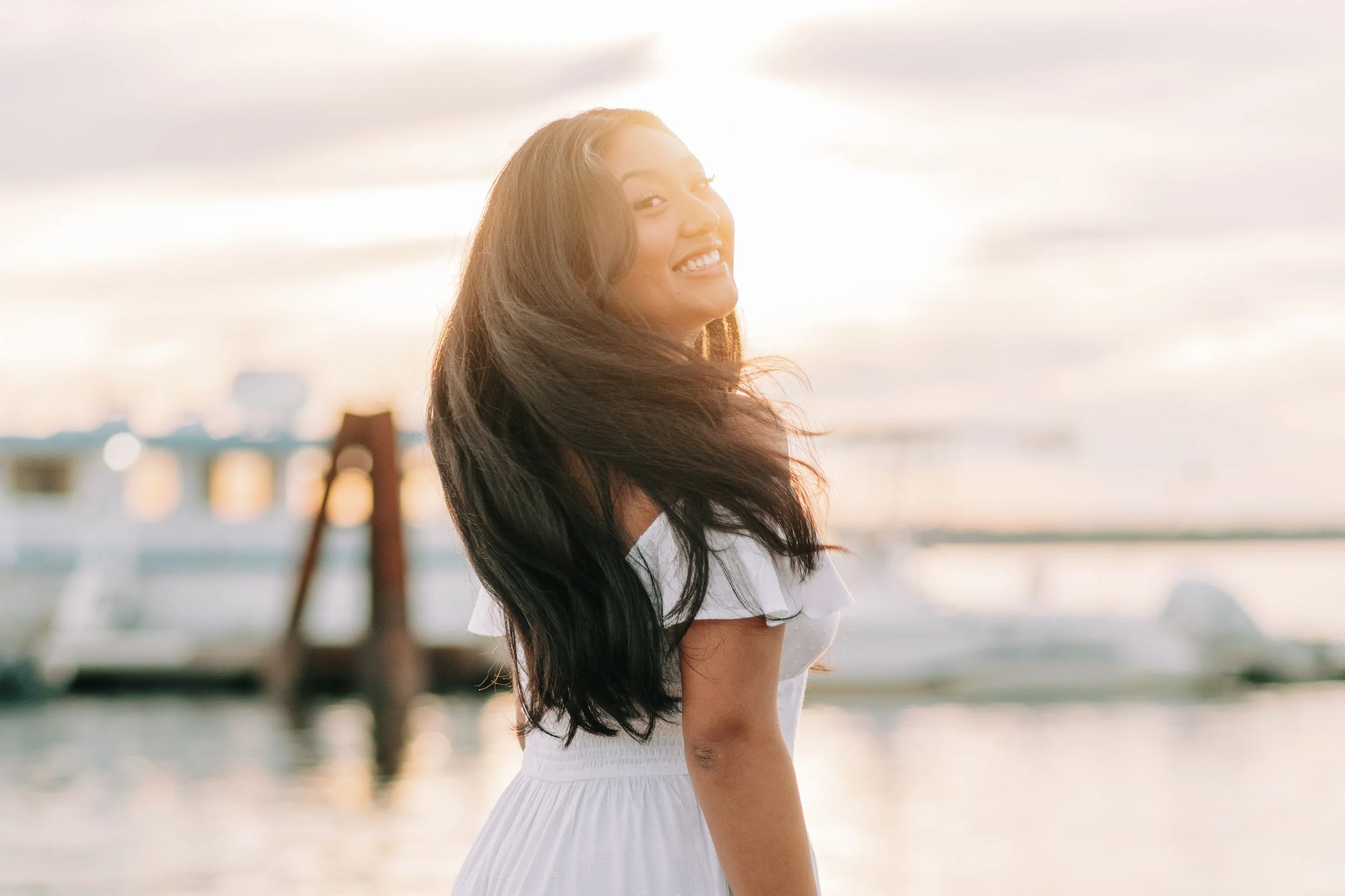 A woman with long dark hair smiling at the camera in a white dress near a waterfront with boats in the background during sunset