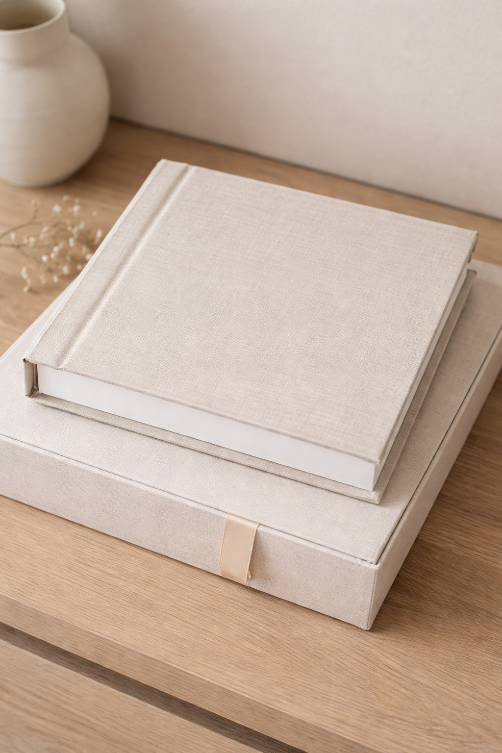 A closed beige fabric-covered photo album on top of a matching box, placed on a wooden surface with a ceramic vase and small dried flowers nearby.