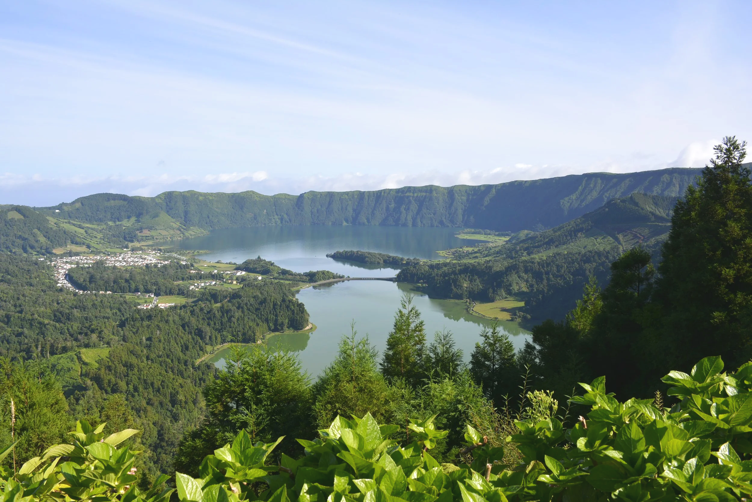 A scenic view of a large lake surrounded by green hills and mountains with a blue sky above.