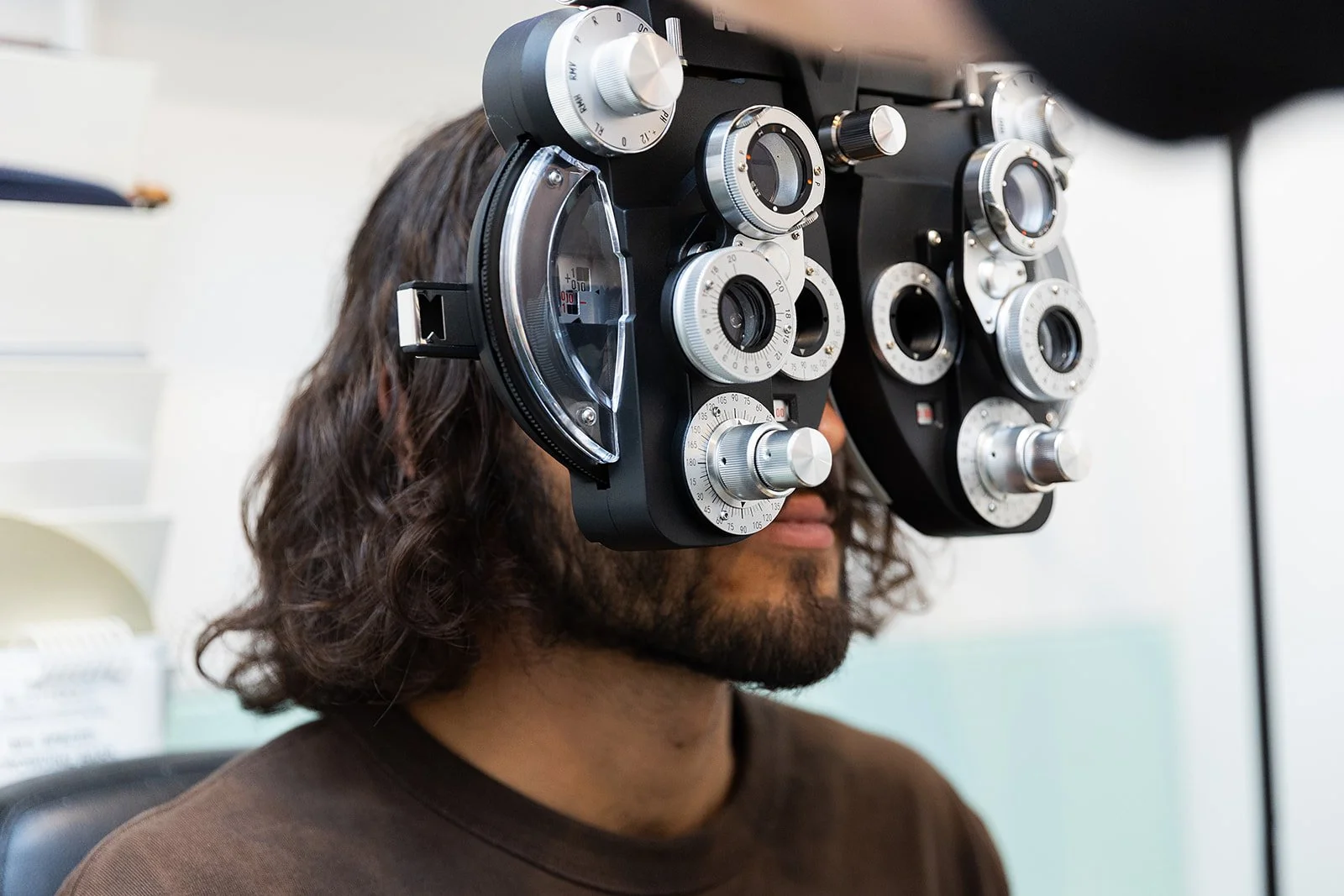 Person with long, wavy hair wearing a phoropter, an eye examination device, during an eye exam.