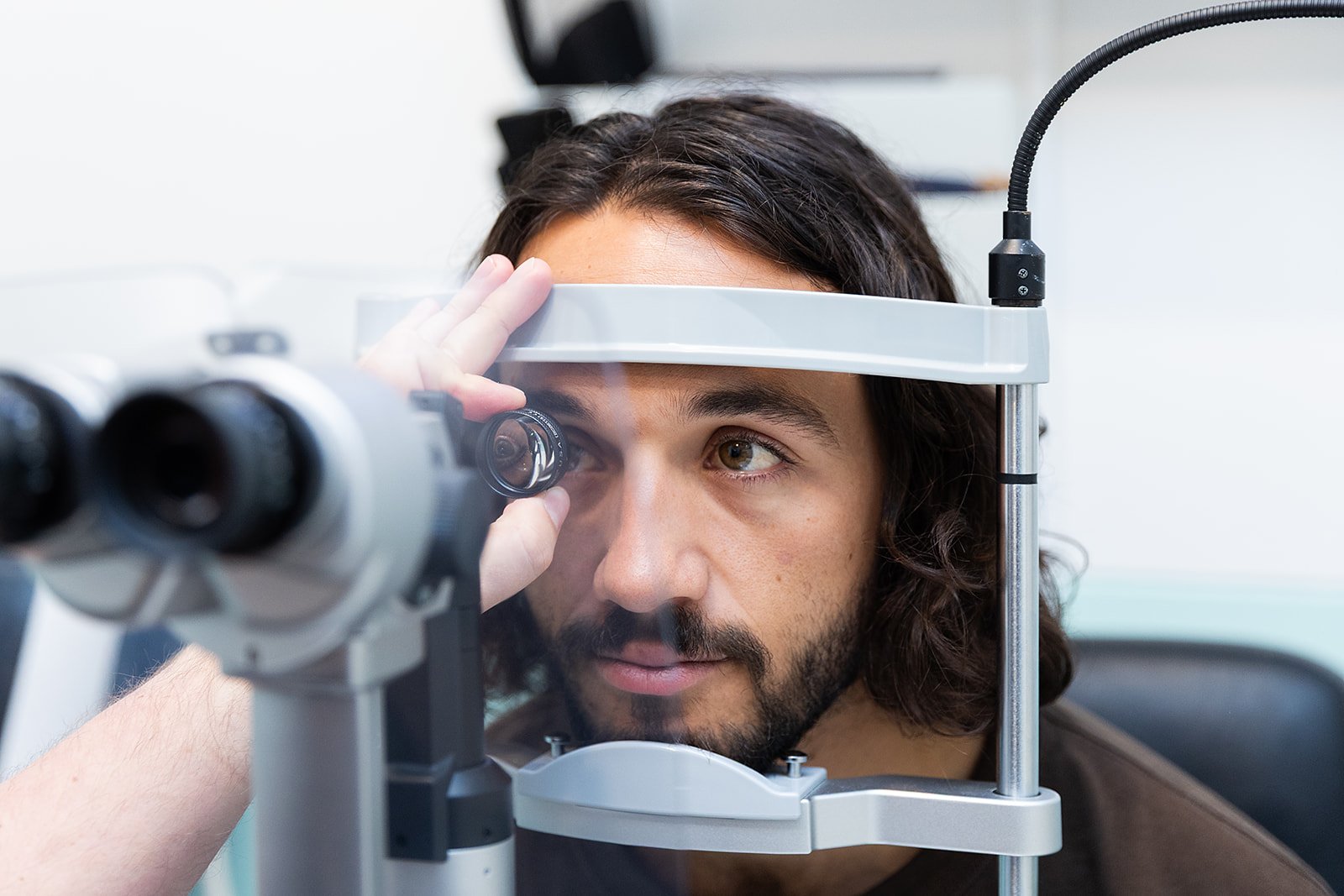 A man with long brown hair and a beard is using a slit lamp for eye examination, looking through a microscope.