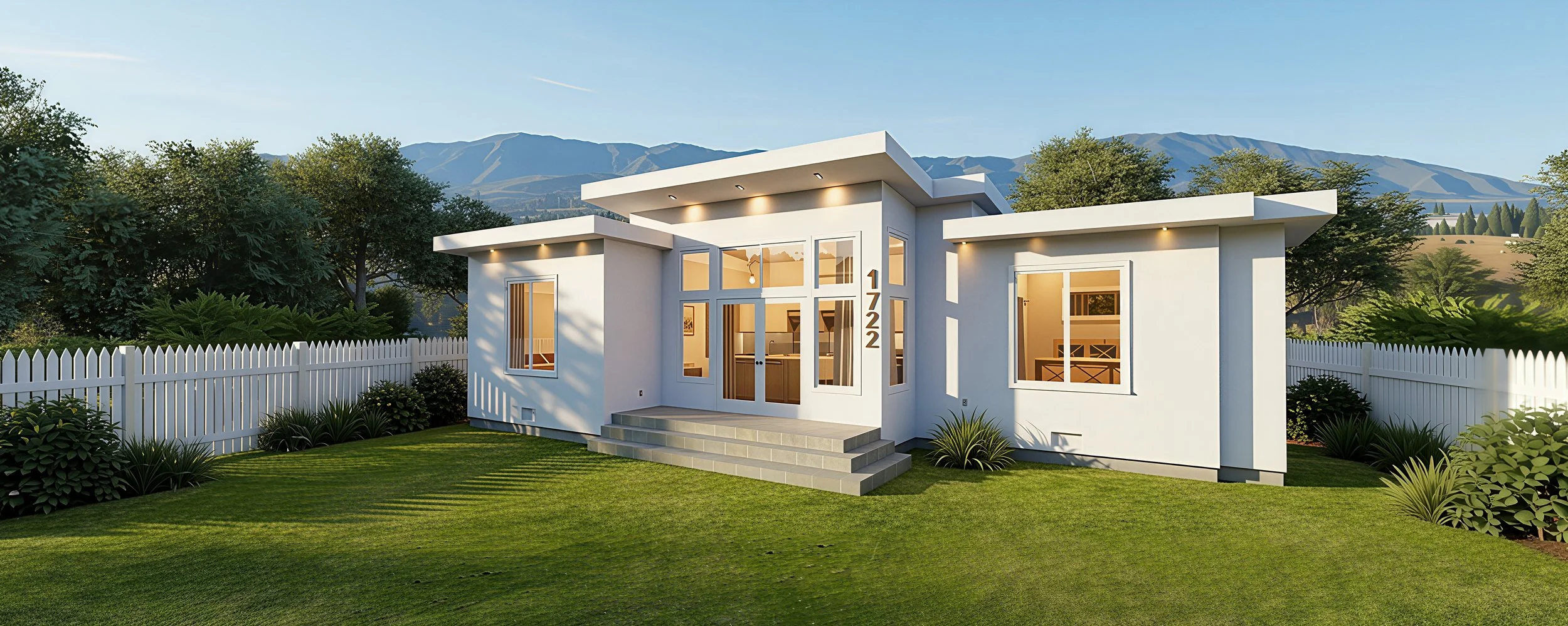 Modern white single-story house with large windows, surrounded by a white picket fence, green lawn, and trees with mountains in the background.