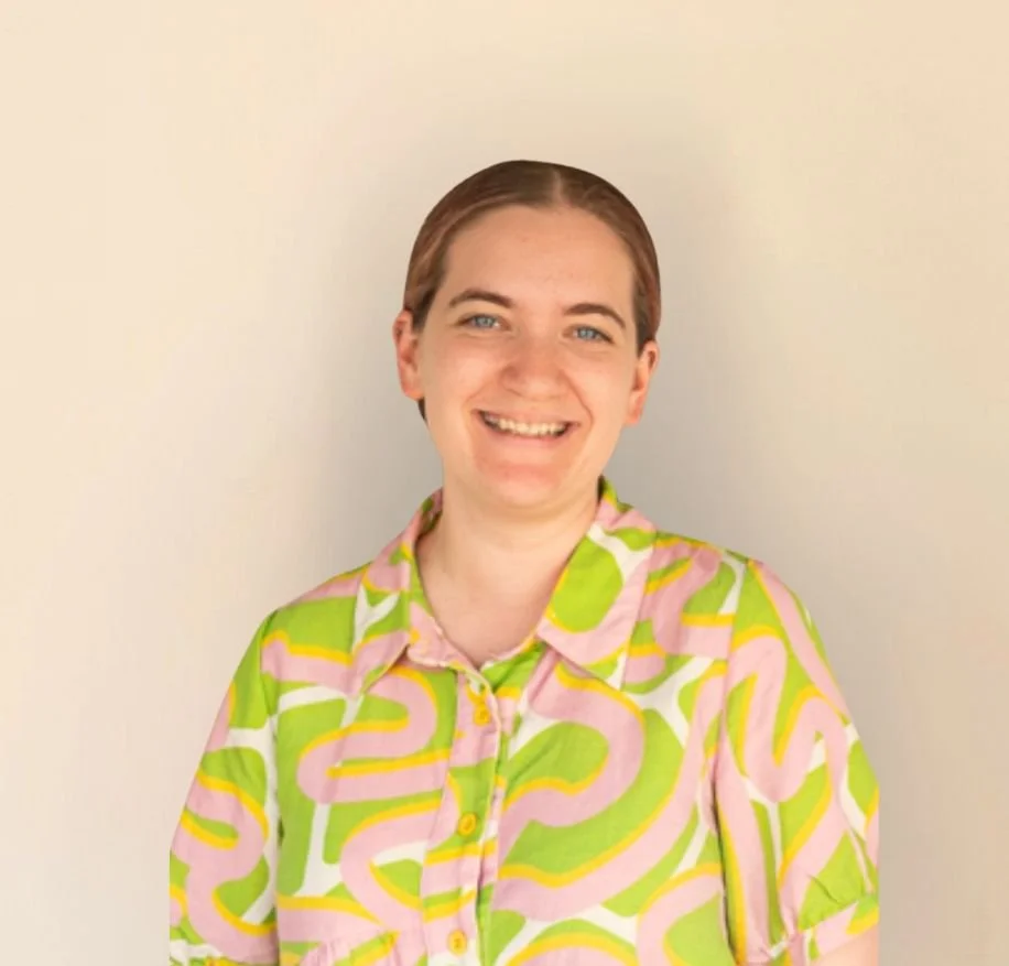 A person with short brown hair and blue eyes, smiling, wearing a colorful pink, green, and yellow patterned shirt, standing against a plain light-colored wall.