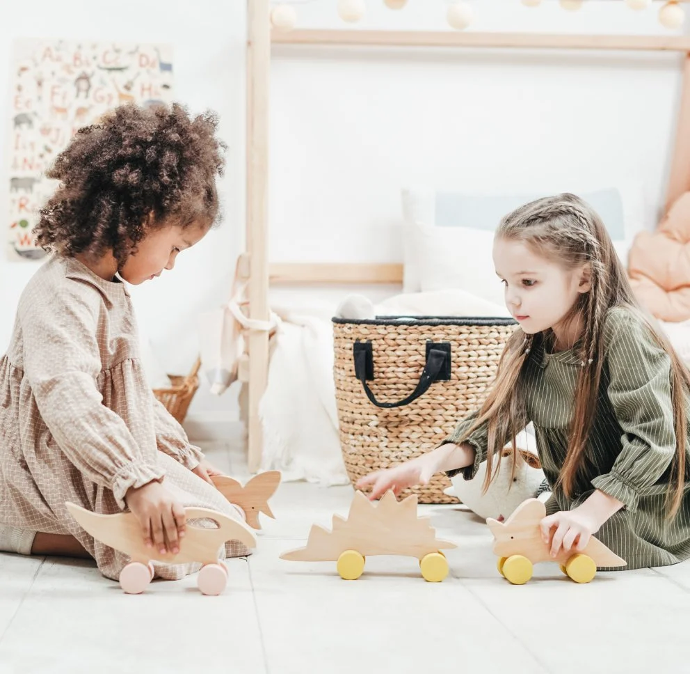 Two young girls playing with wooden toy vehicles shaped like animals on wheels in a bright room.