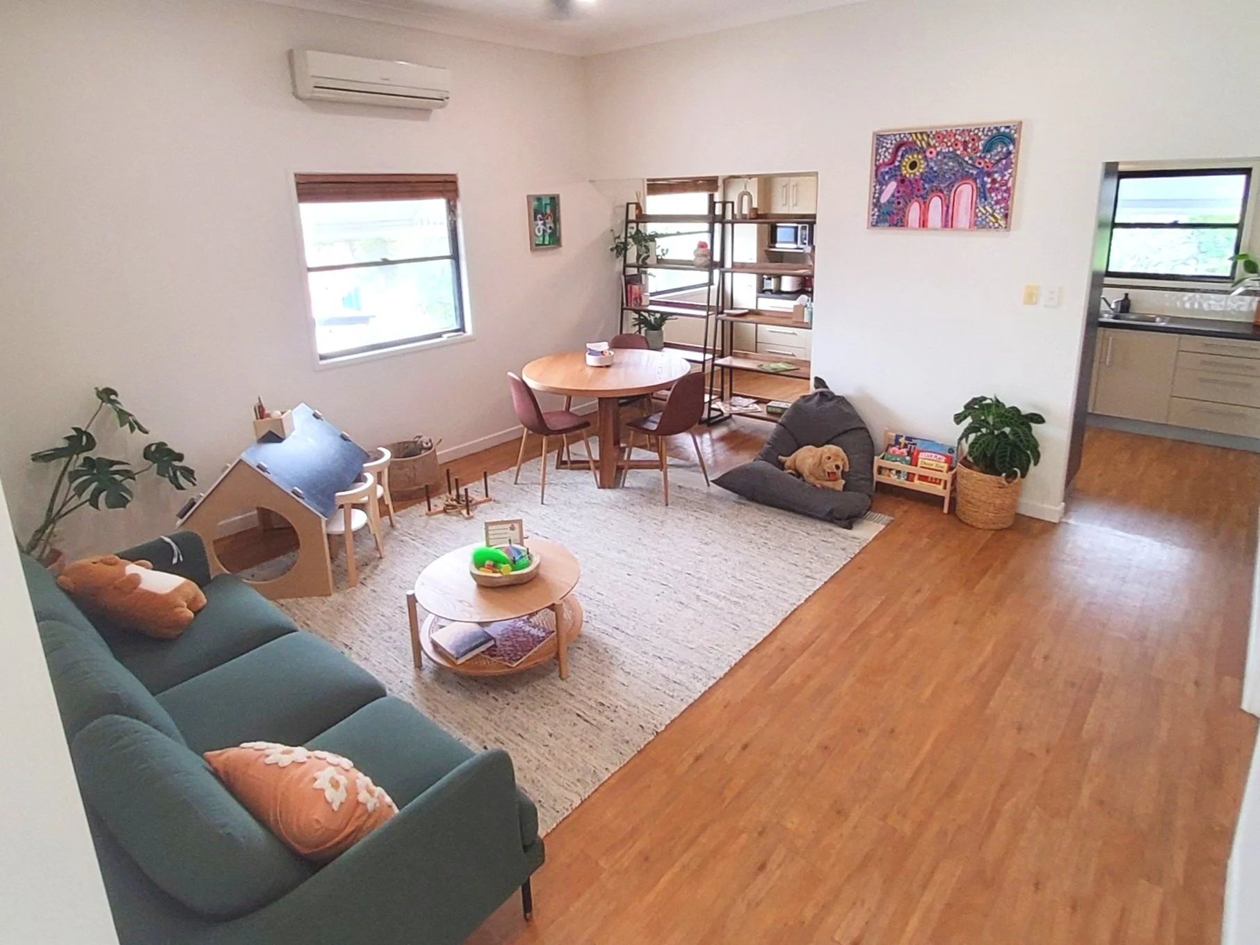 A cozy living room with a green sofa adorned with orange and floral pillows, a round wooden coffee table with a tray of toys and a book, a beige area rug, a dark gray pet bed with a happy golden retriever, a bookshelf, a round wooden dining table with six pink chairs, art on the walls, and a view into the kitchen through an open doorway. The room has hardwood floors, windows letting in natural light, and a white air conditioning unit on the wall.