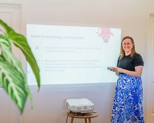 A woman smiling and holding a remote, giving a presentation on emotional regulation with a slide projected on a white wall, in a room with a potted plant in the foreground.