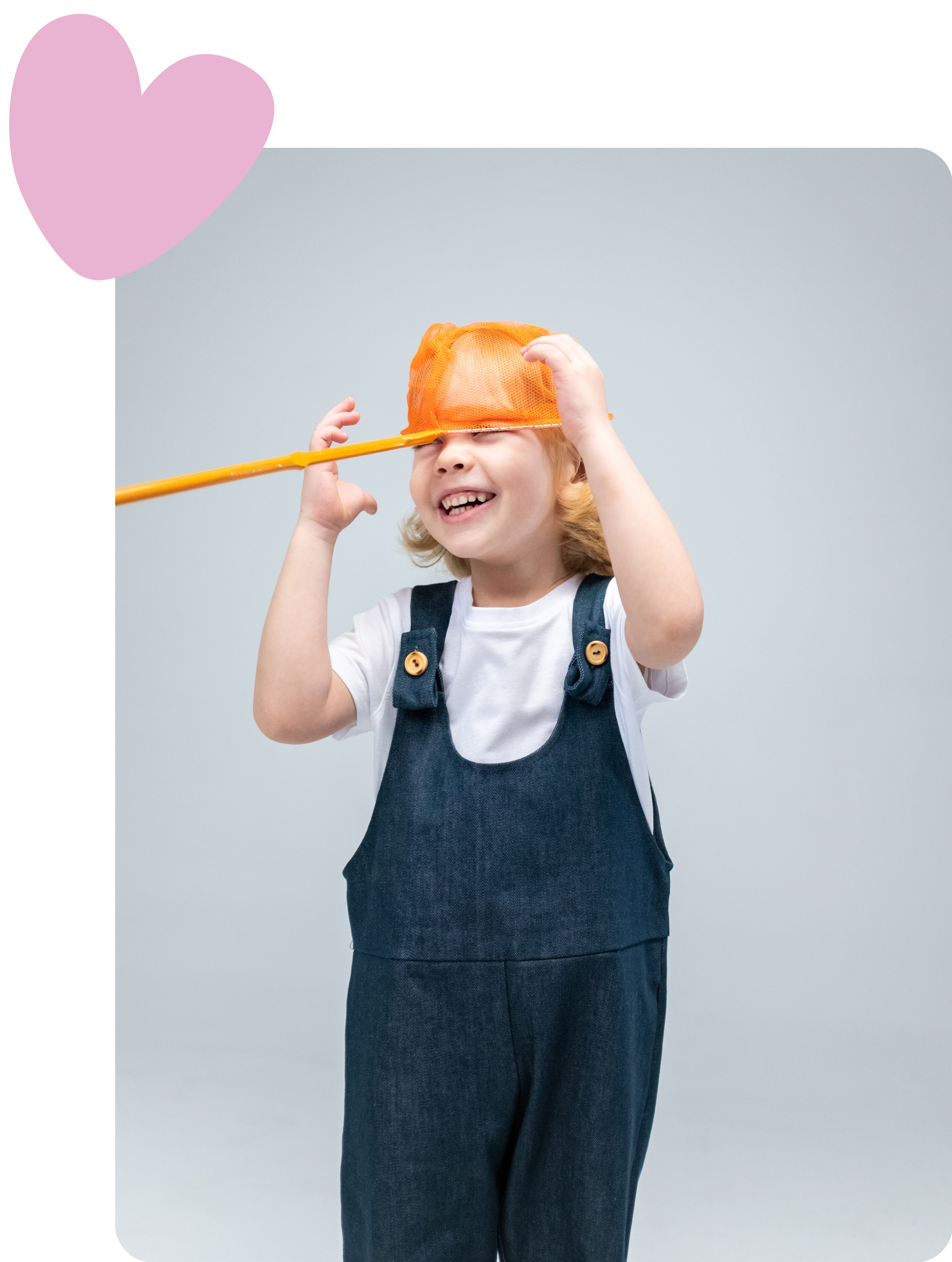 A young child with blonde hair smiling and holding an orange mesh toy over their eyes, wearing denim overalls and a white shirt, against a plain gray background.