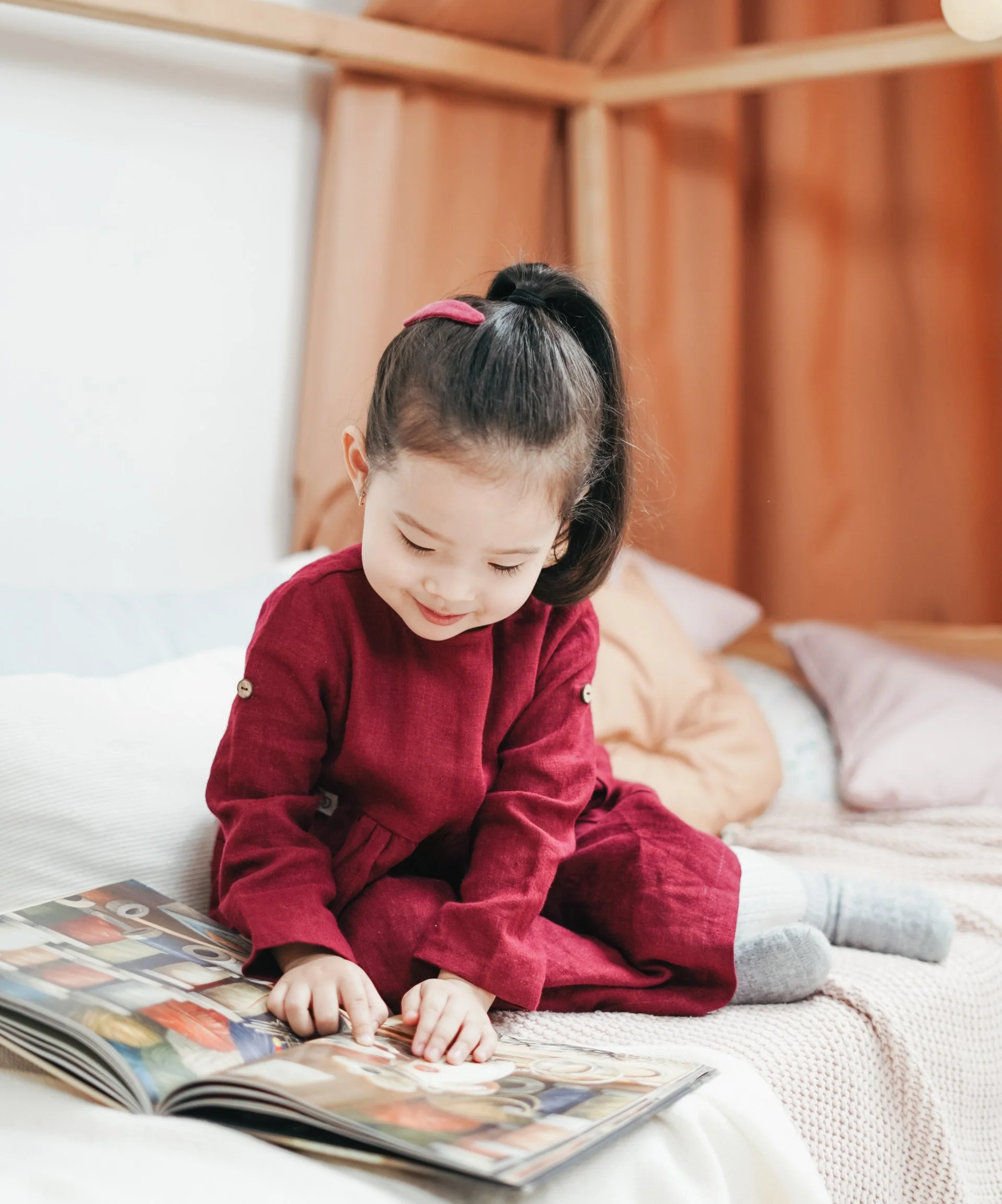 A young girl with dark hair tied in a ponytail, wearing a red velvet dress, is sitting on a bed and looking at a colorful magazine spread out on the bed. She is focused on the magazine, with her hands pointing at something in the magazine, and the background features wooden wall panels.