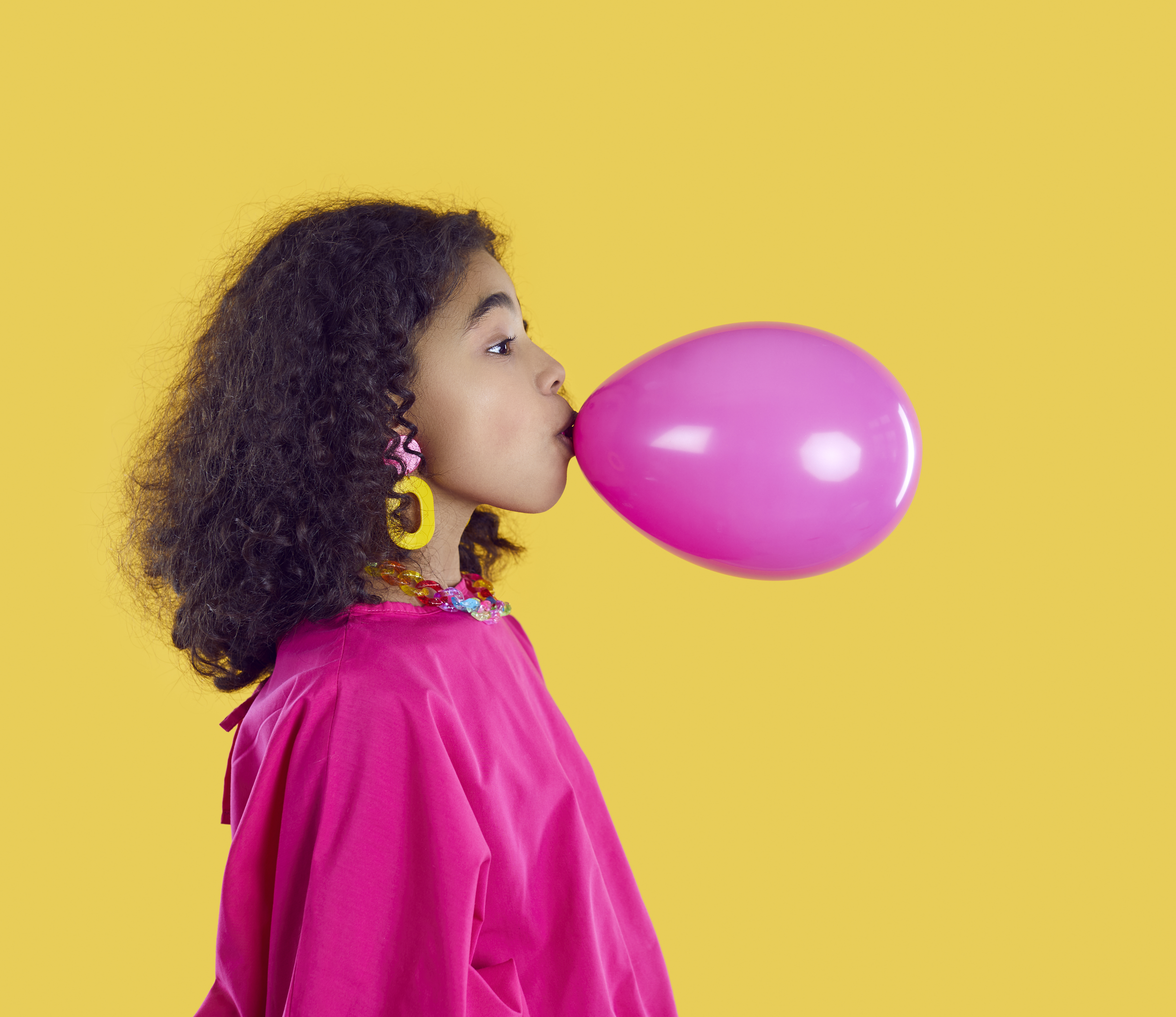 A young girl with curly hair and yellow earrings is blowing up a pink balloon against a yellow background.
