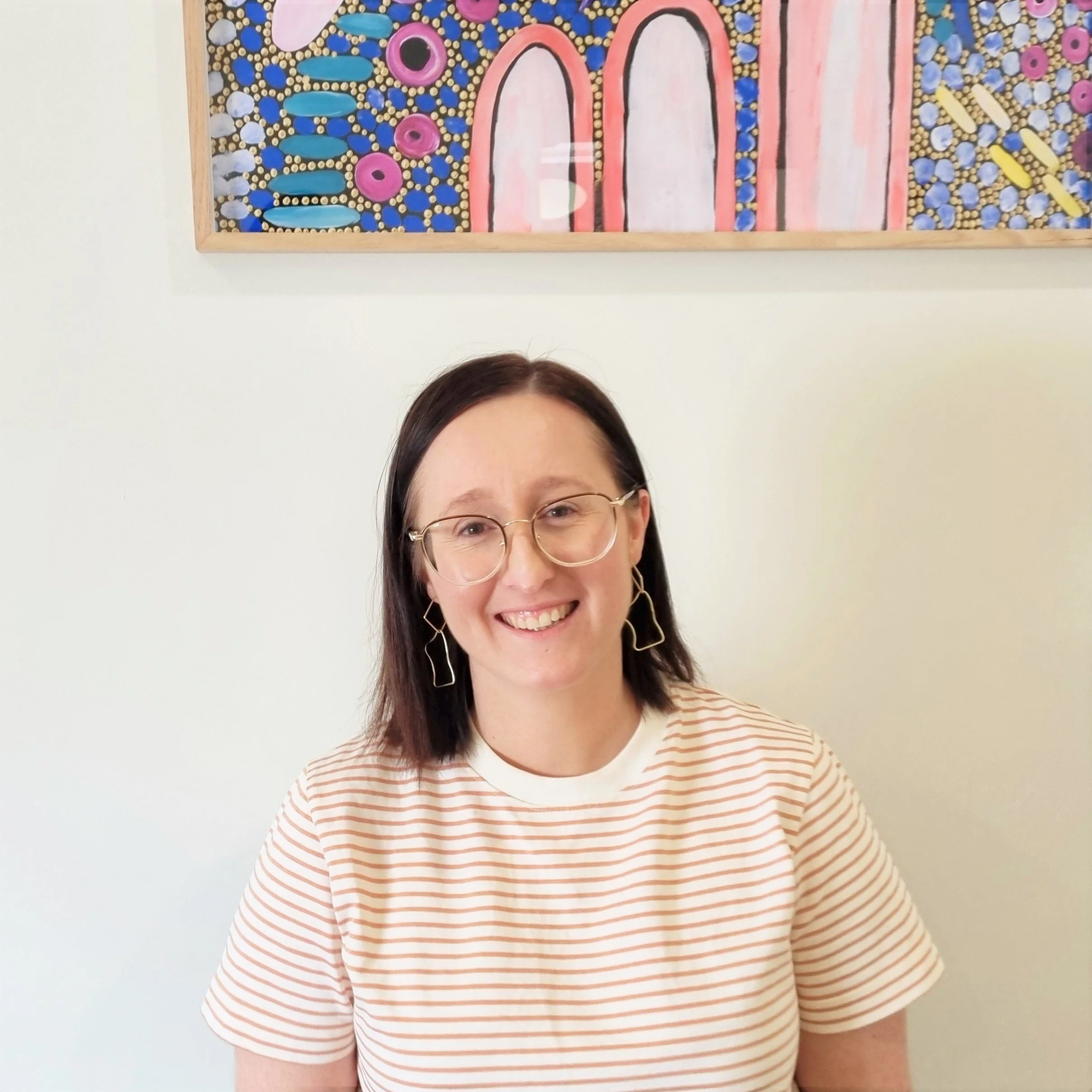 A woman smiling, wearing glasses and earrings, with a striped shirt, standing in front of a white wall with a colorful abstract painting hanging above her.