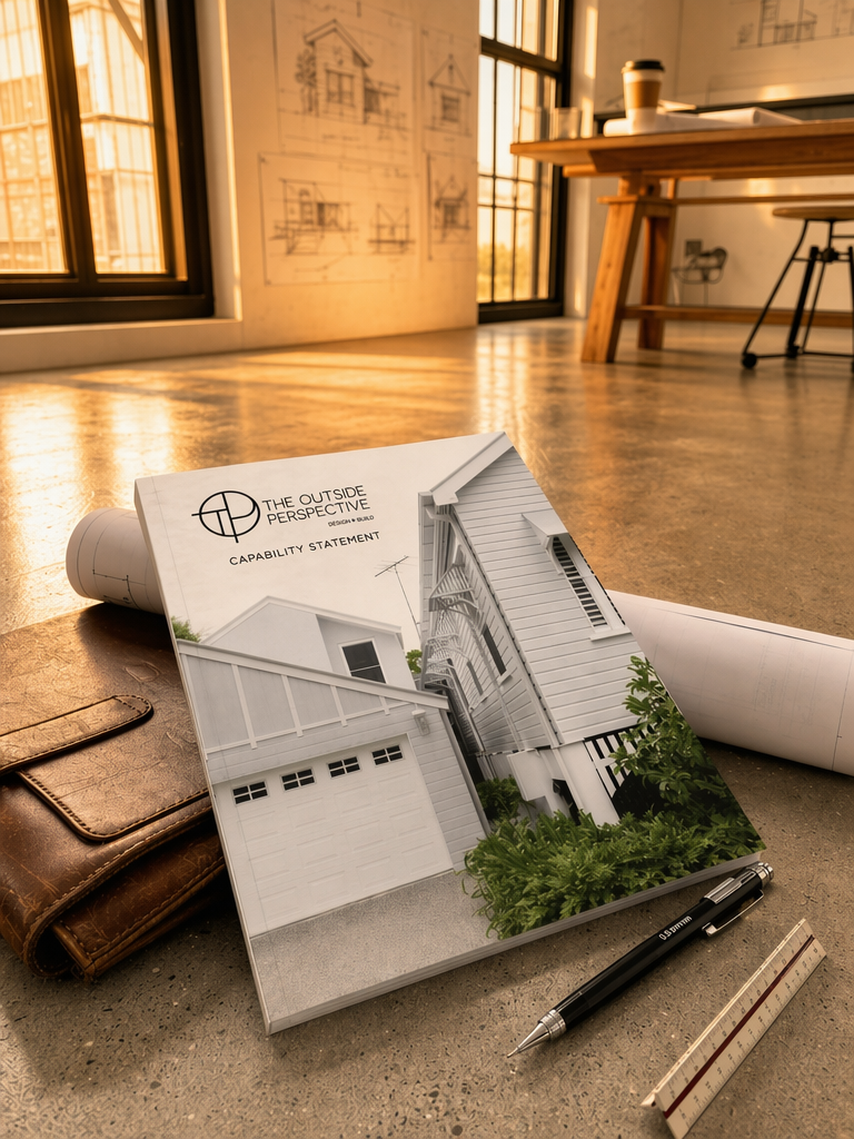 A brochure titled 'The Outside Perspective' featuring a modern white house with a garage and greenery, placed on a concrete floor with a pen, ruler, brown leather wallet, and architectural rolled blueprints nearby, in a bright room with wooden furniture and large windows.