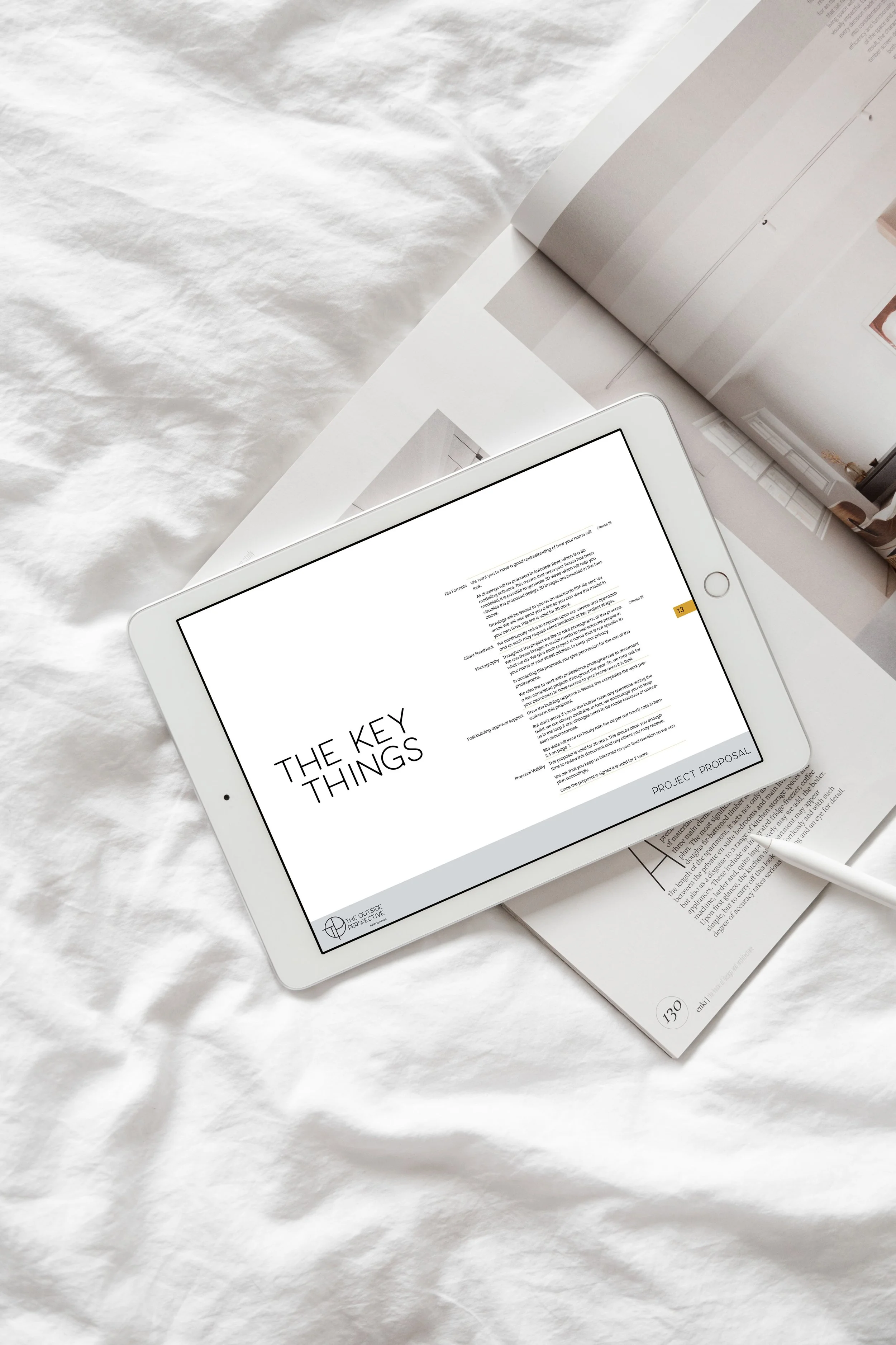 An electronic tablet displaying a document titled 'The Key Things' with the project proposal at the bottom right. The tablet rests on open magazines or catalogues on a white wrinkled fabric surface.