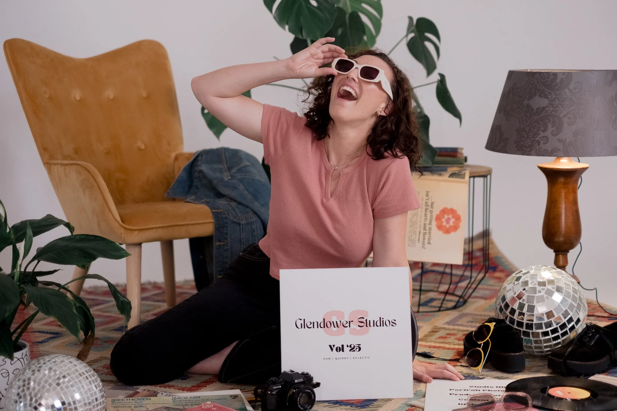 A woman with curly hair wearing sunglasses and a pink shirt, laughing and leaning back on a couch in a decorated indoor space with plants, a lamp, and some jewelry items, at Glendower Studios event.