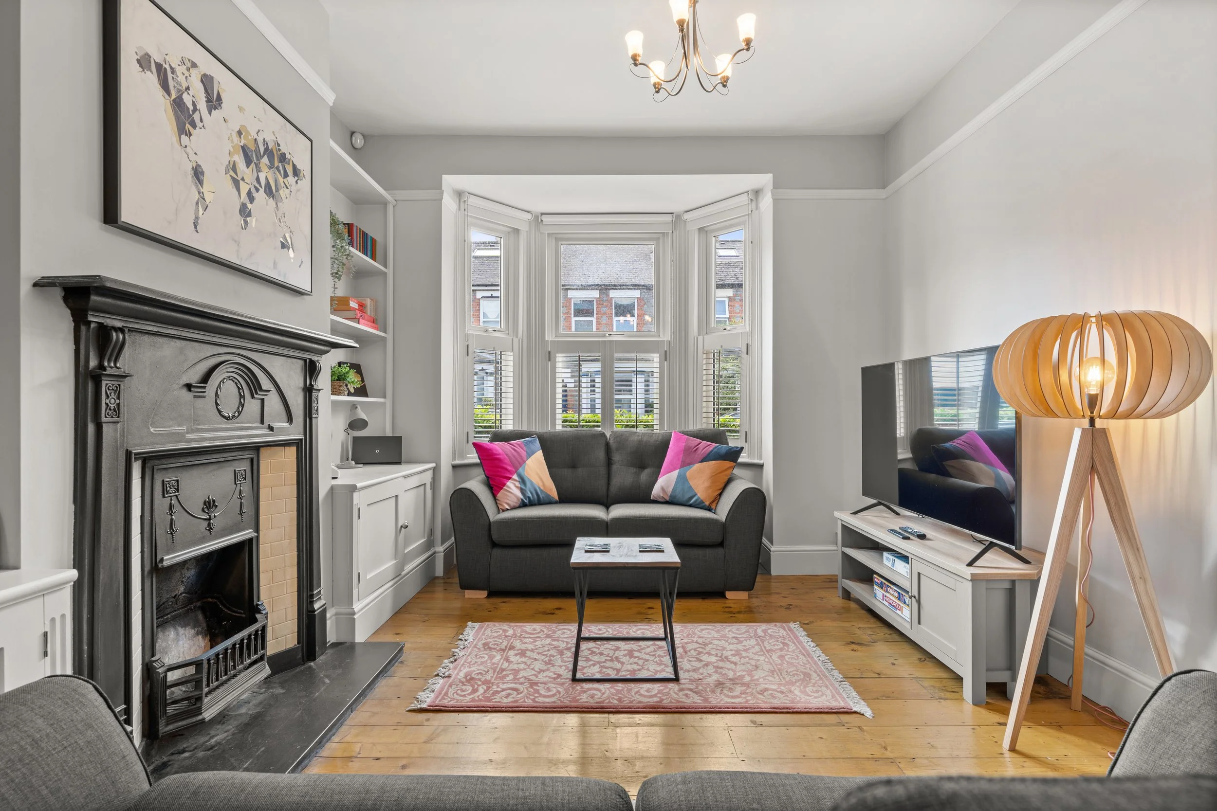 Living room with a gray sofa, decorative pillows, a fireplace, a flat-screen TV, a floor lamp, a pink area rug, a window with shutters, and a chandelier.