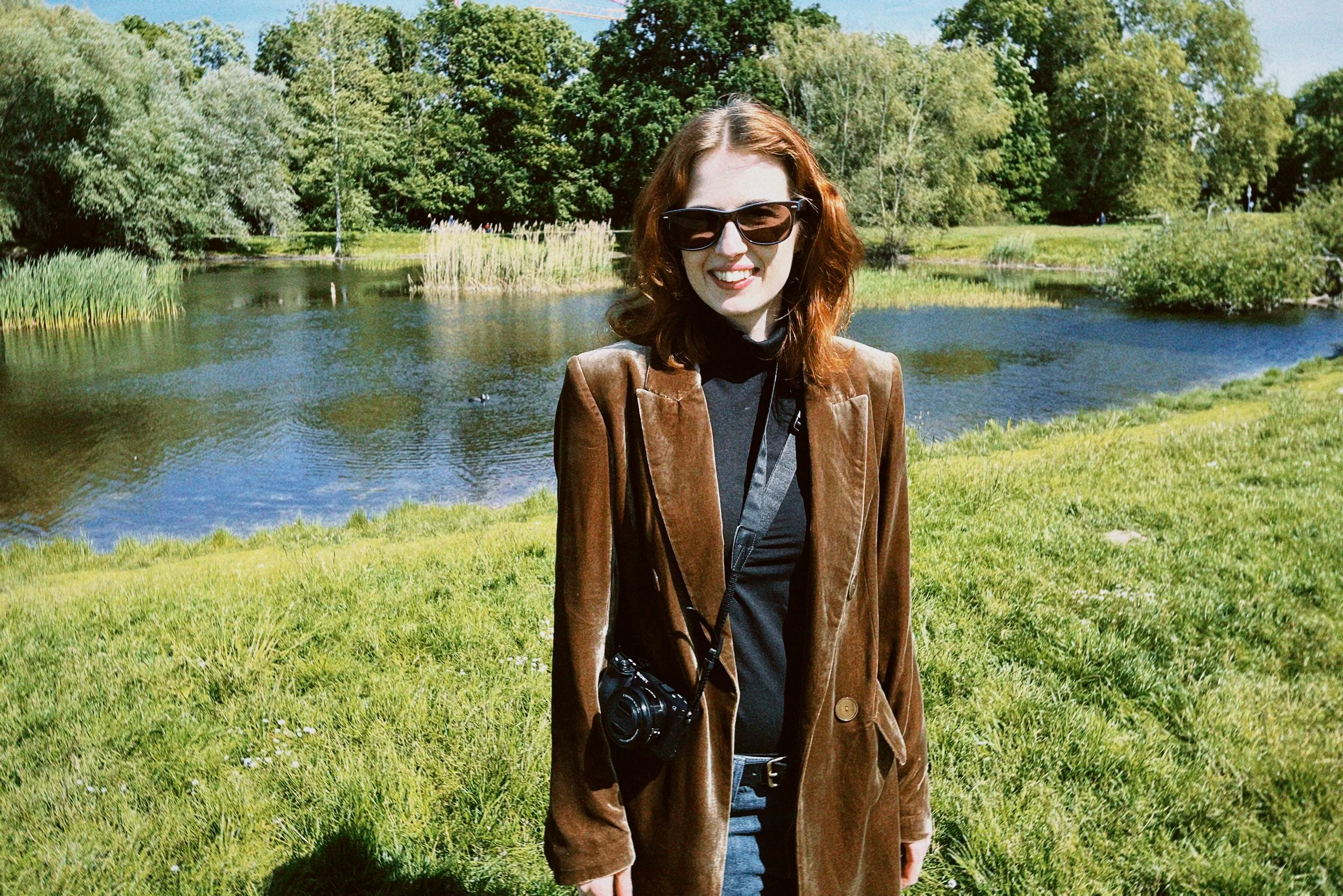 A woman with curly red hair wearing sunglasses, a brown jacket, and a camera hanging around her neck, standing in front of a pond with green trees in the background on a sunny day.