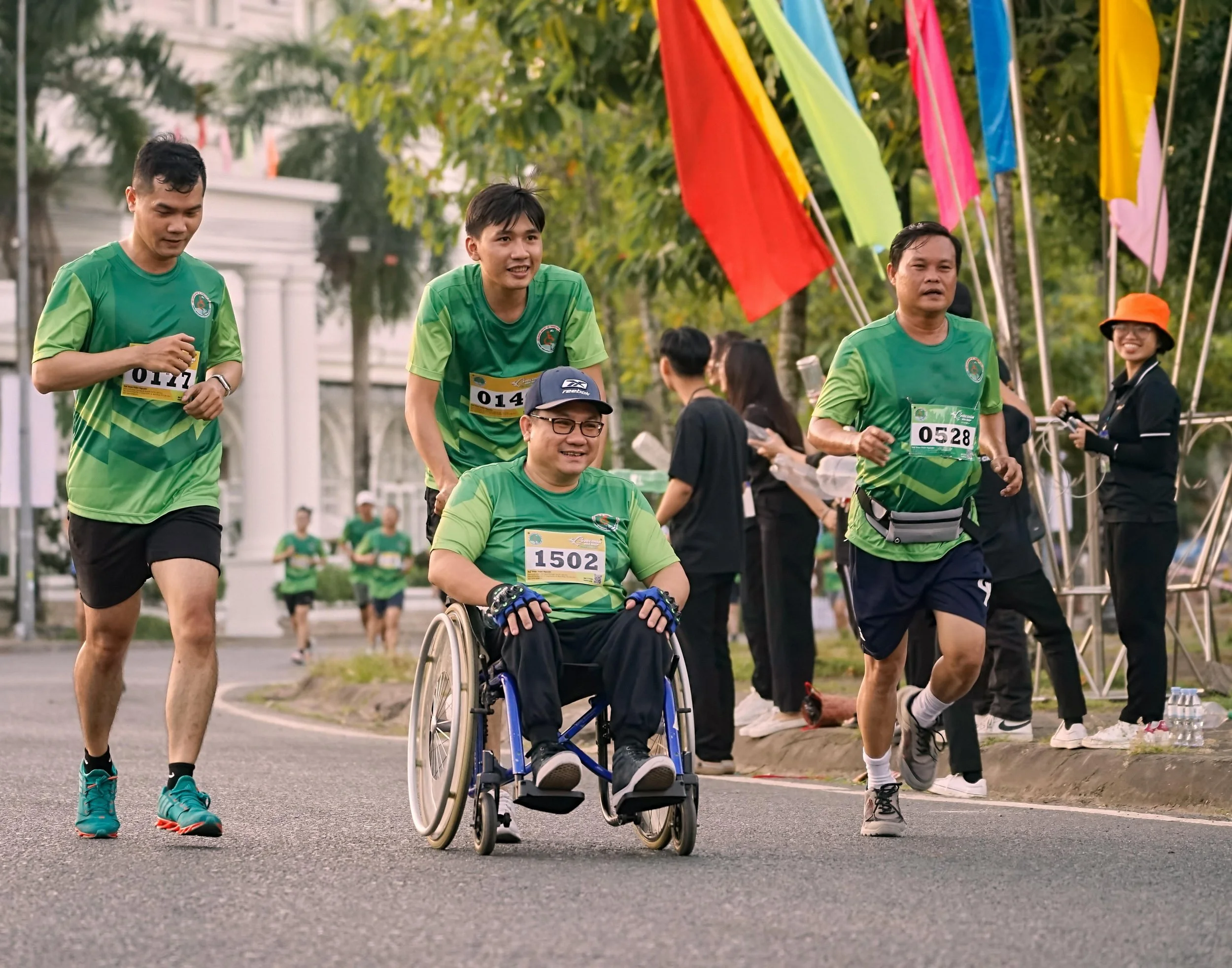 Group of people running in a race, including a man in a wheelchair, wearing green shirts and bib numbers, along with others running beside him, outdoors with colorful flags in the background.