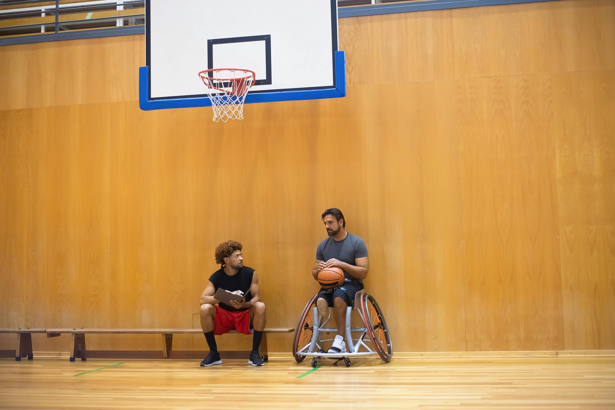Two men talking in a gymnasium with a basketball hoop and a wooden wall. One man is sitting on a bench holding a clipboard, wearing athletic gear. The other man is in a wheelchair, holding a basketball, and is engaged in conversation.