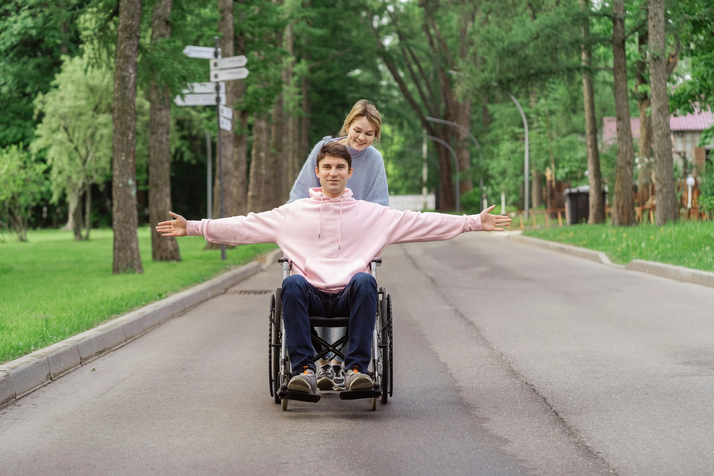 A young man in a pink hoodie sitting in a wheelchair with arms outstretched, and a young woman standing behind him, smiling, in a park with trees lining the pathway.