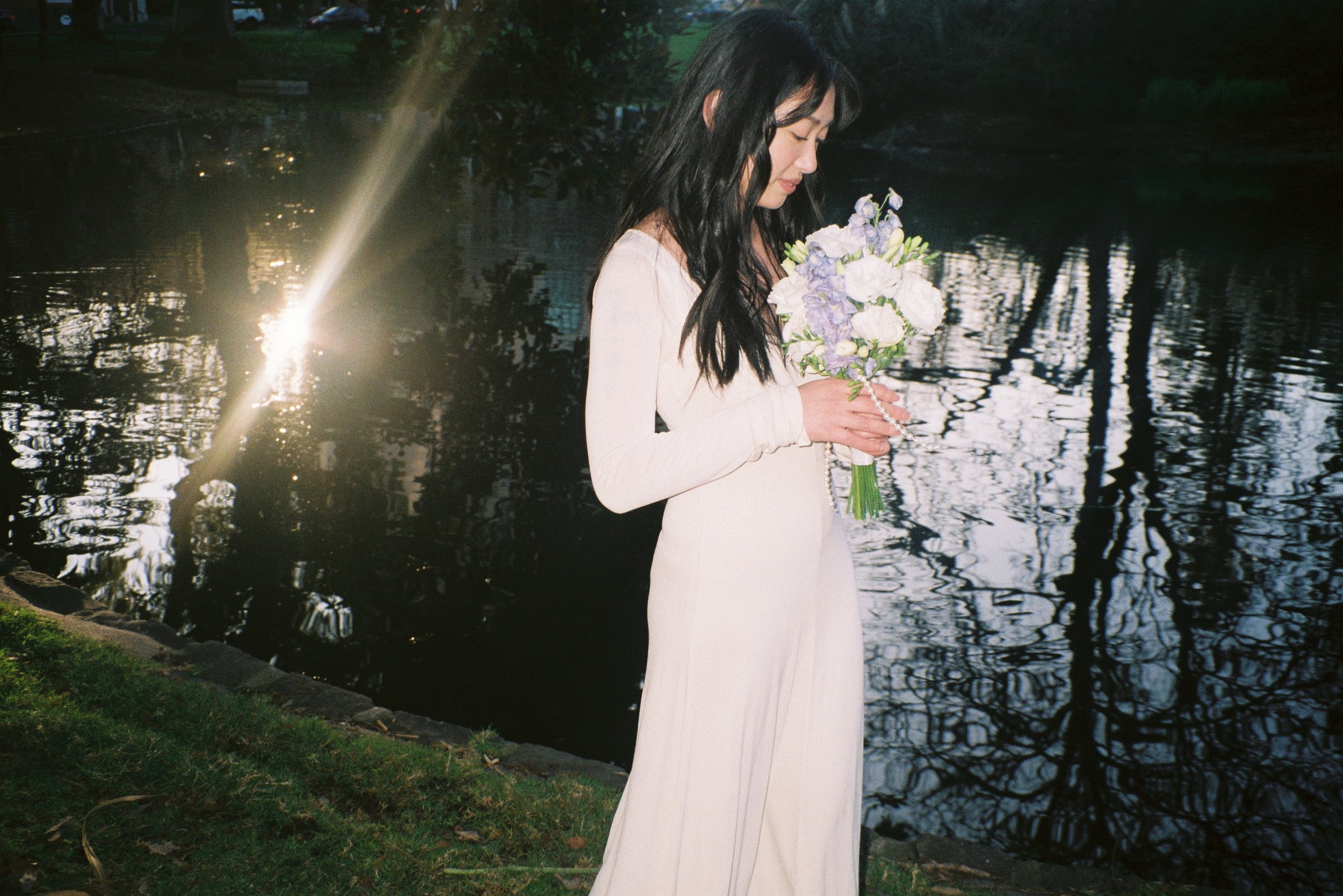 A woman in a long white dress holding a bouquet of flowers stands by a body of water in a park during the evening, with trees reflected on the water's surface.
