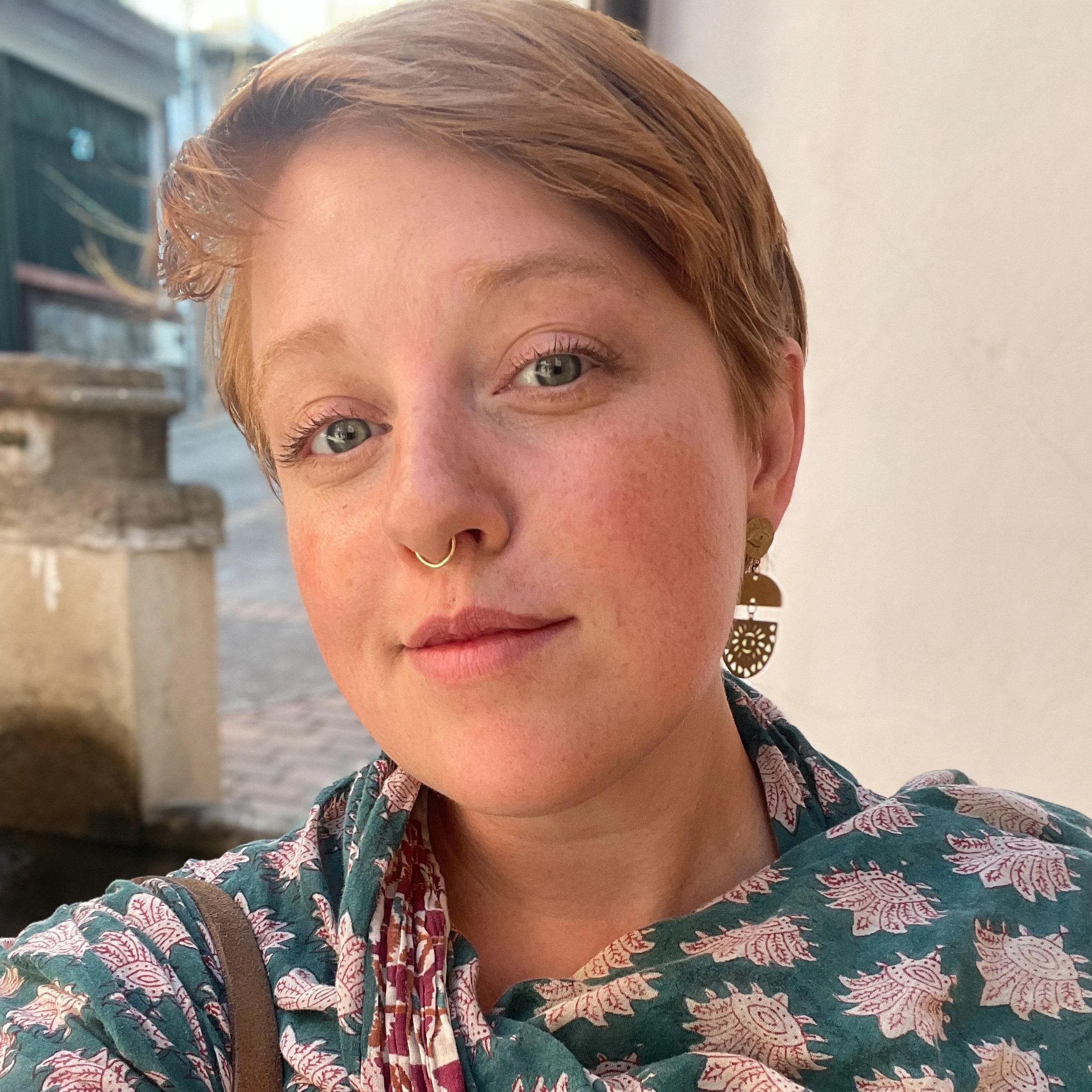 A photo of Katie Kaestner, a white Jewish person with short red hair and brown glasses wearing a dark blue shirt.