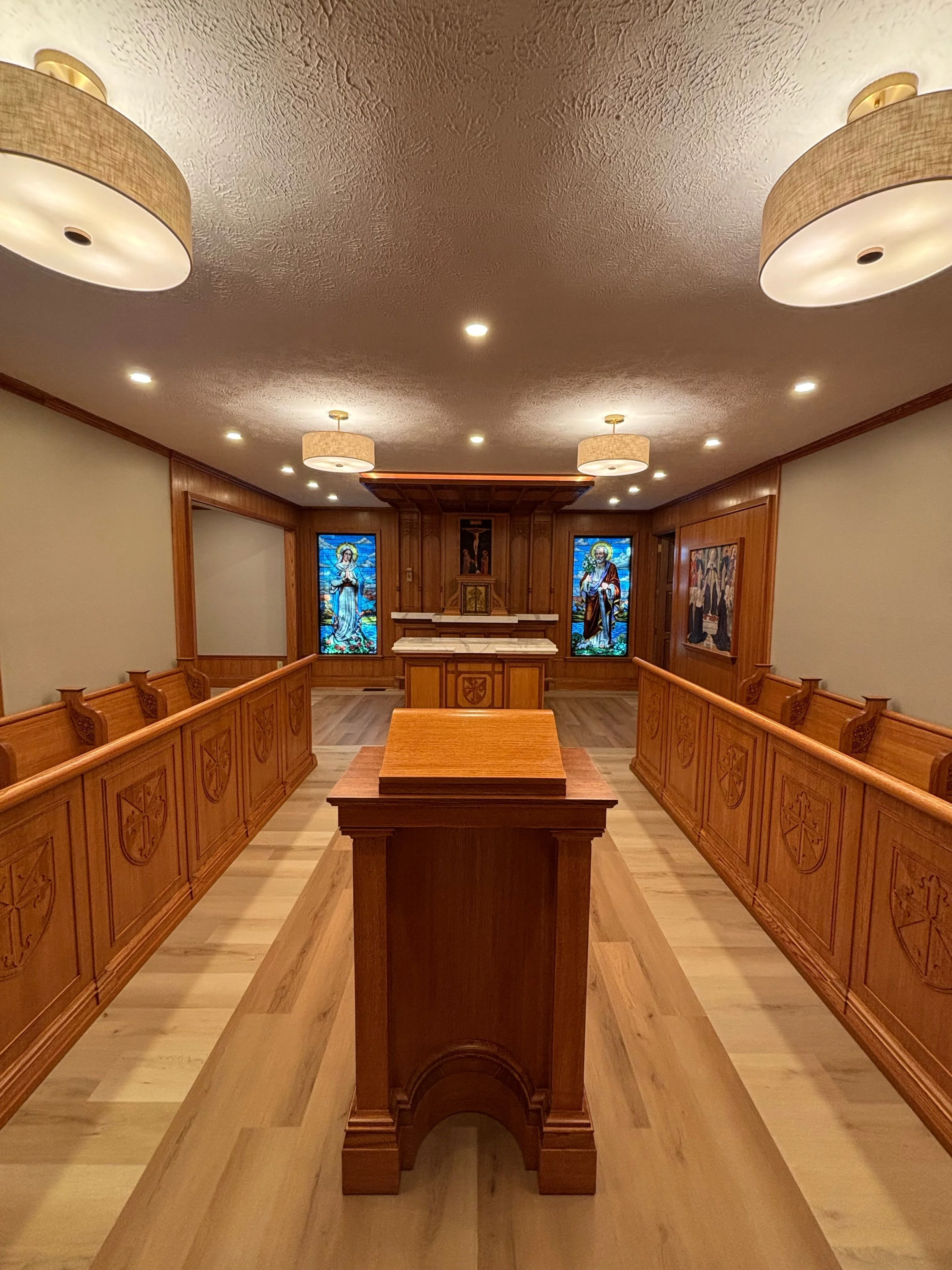 The interior of a small chapel with wooden pews, stained glass windows depicting religious figures, a wooden altar, and a ceiling with chandeliers and recessed lighting.
