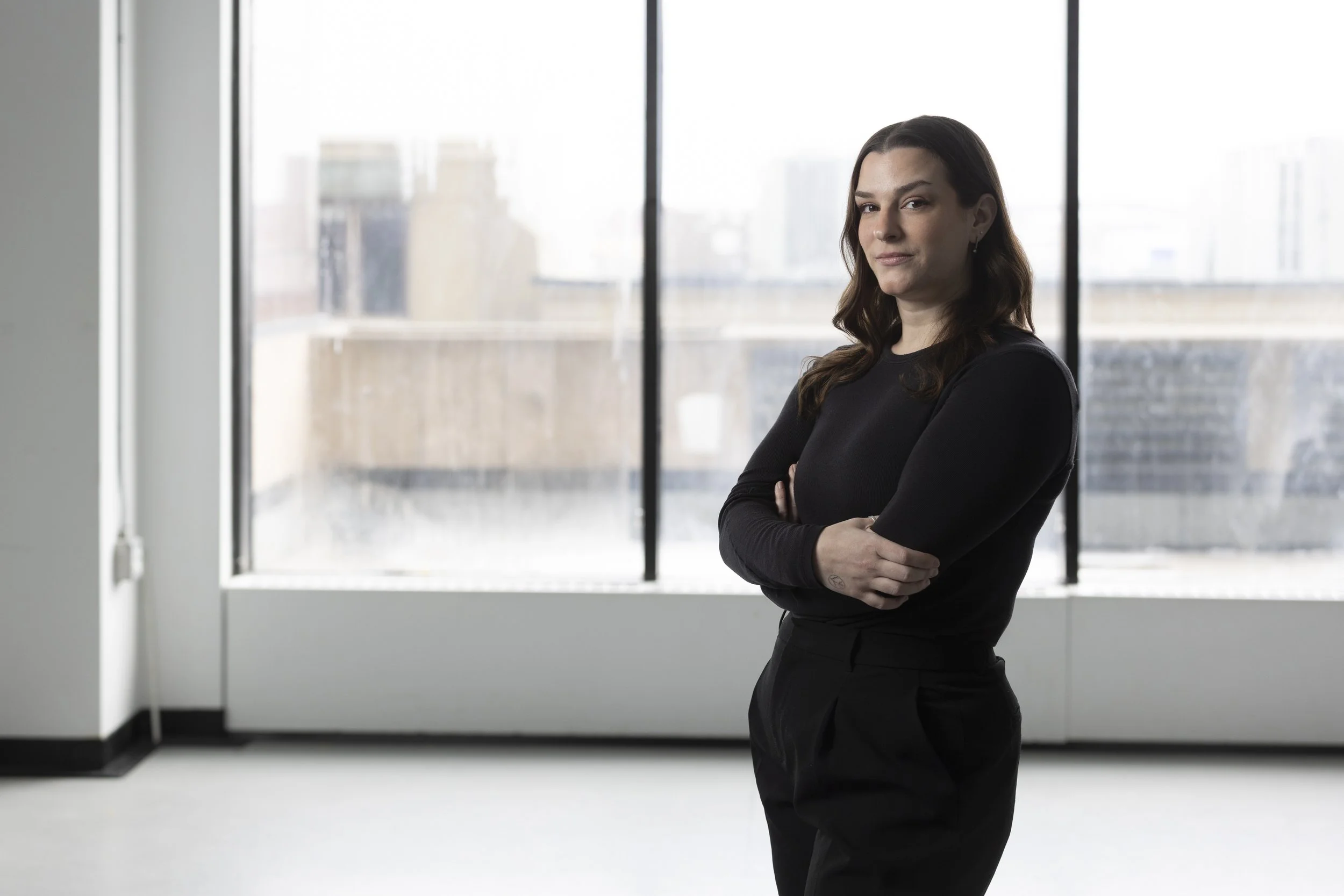 A woman with dark brown hair wearing a black long-sleeve shirt and black pants standing with arms crossed in a modern room with large window overlooking city buildings.