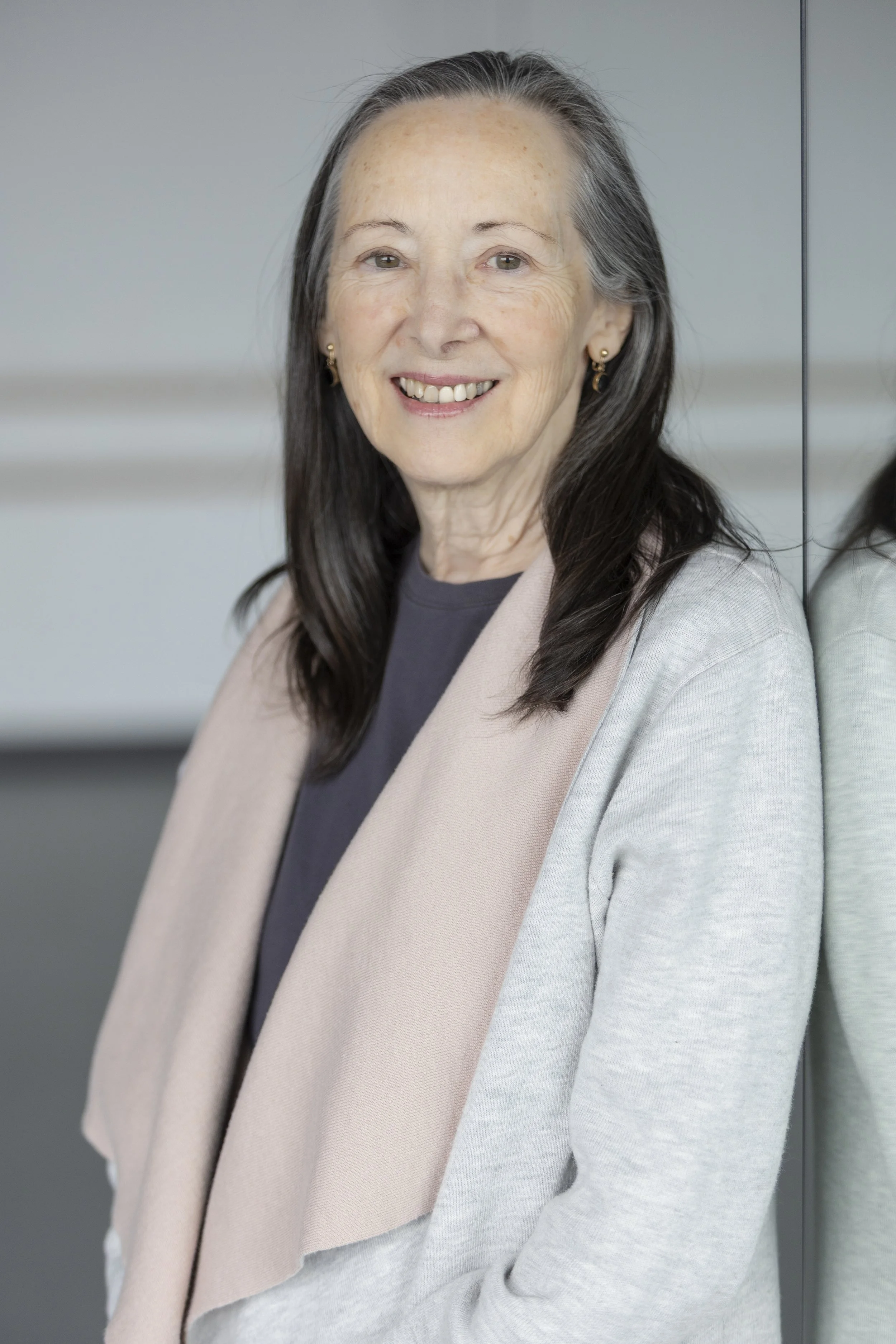 A smiling woman with long gray and brown hair standing next to a mirror in a modern office setting, wearing a beige blazer and dark shirt.