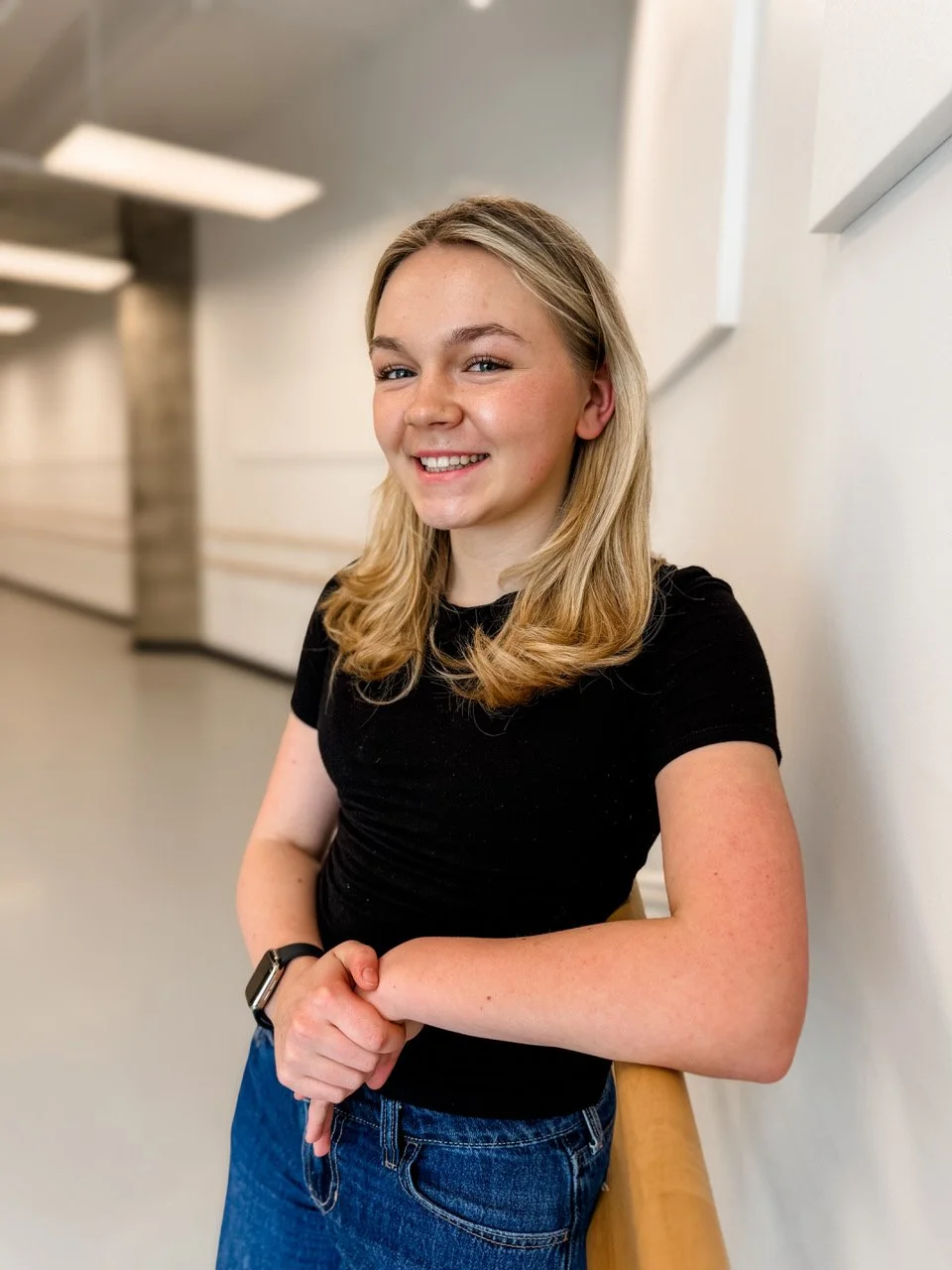 Young woman with blonde hair smiling, wearing a black t-shirt, blue jeans, and a smartwatch, standing in a hallway.