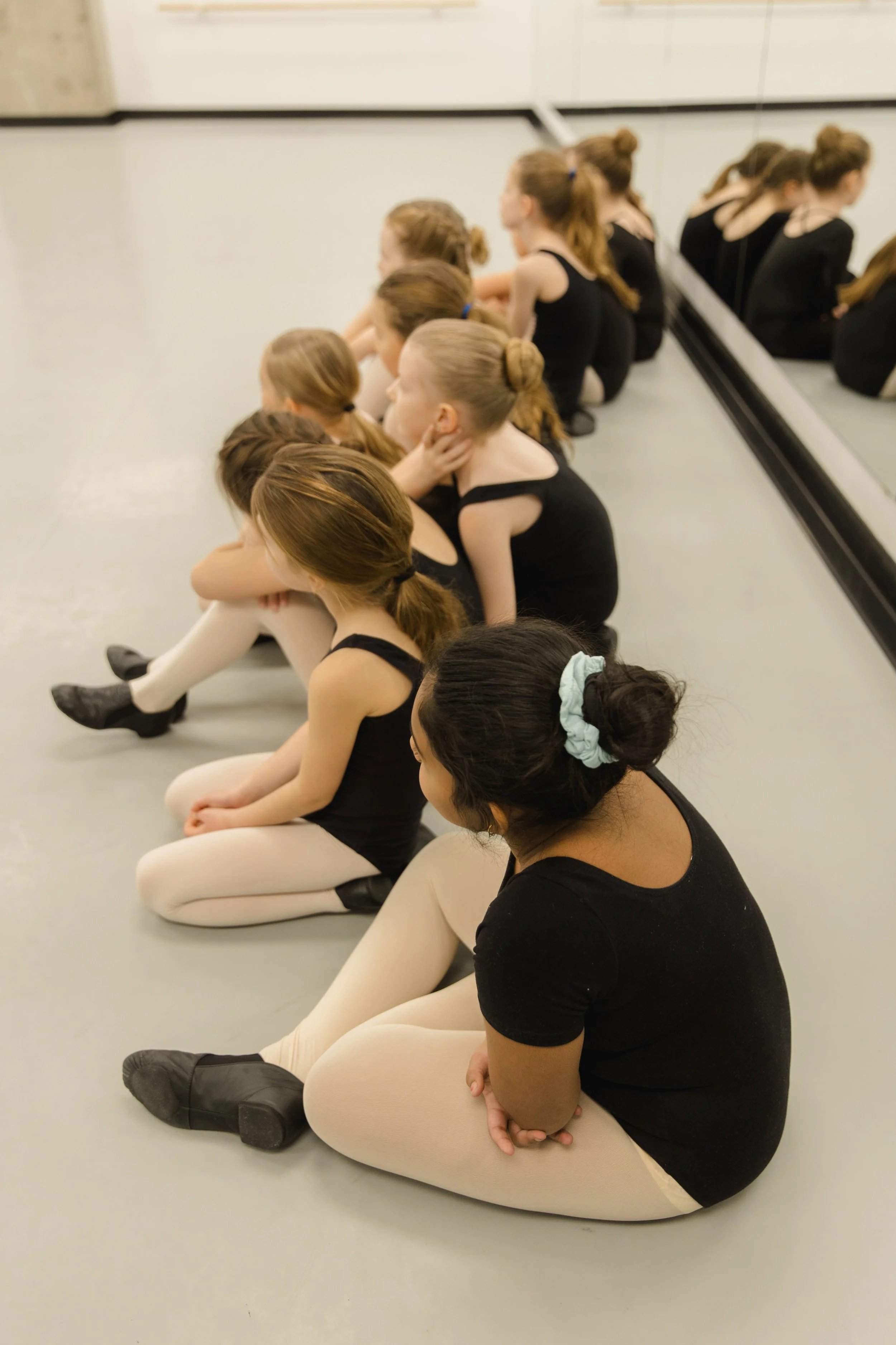Group of young ballerinas sitting on the floor in ballet attire, facing away in a dance studio.