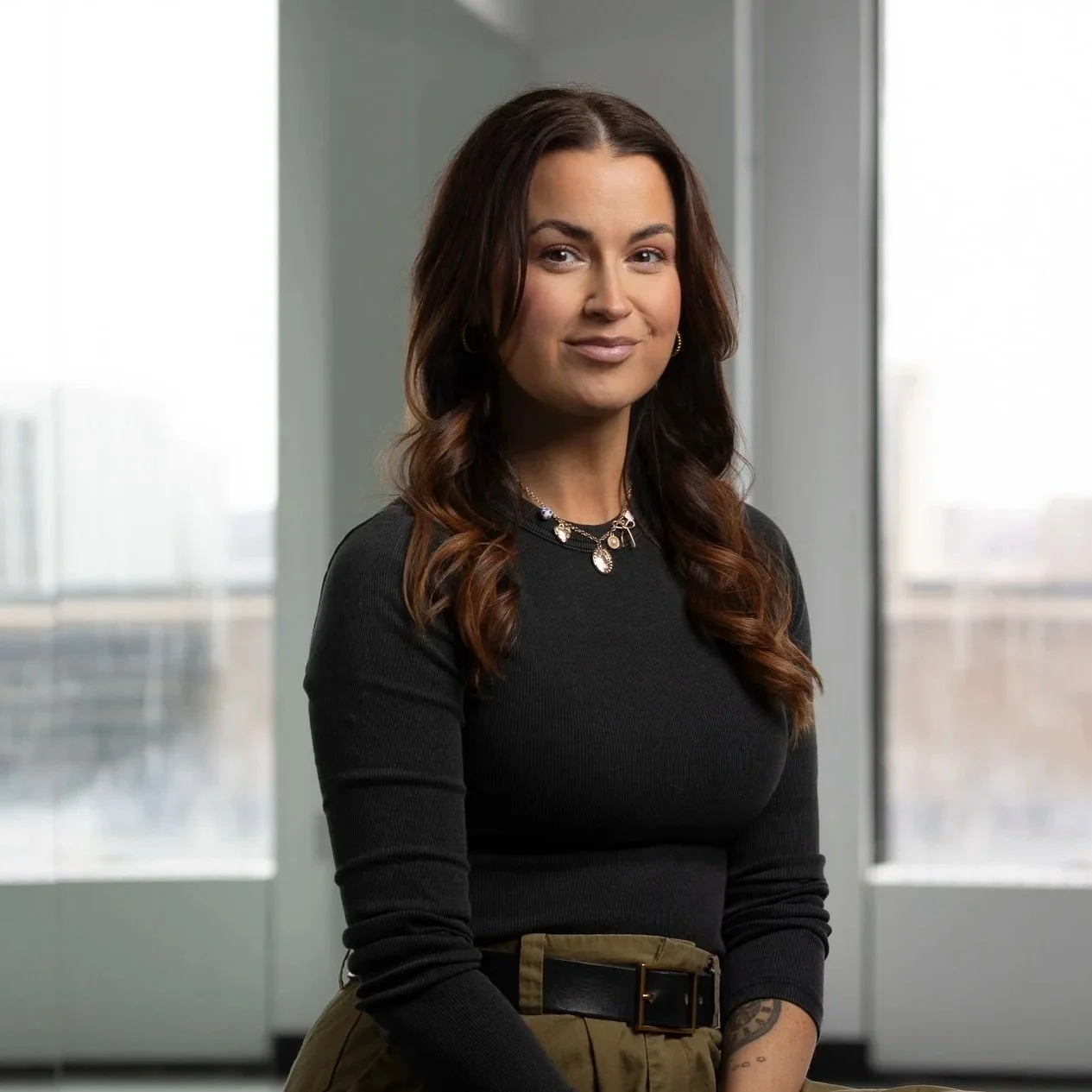 A woman with long brown hair and light skin, wearing a black long-sleeve top and khaki pants, sitting in front of large windows with a cityscape view.