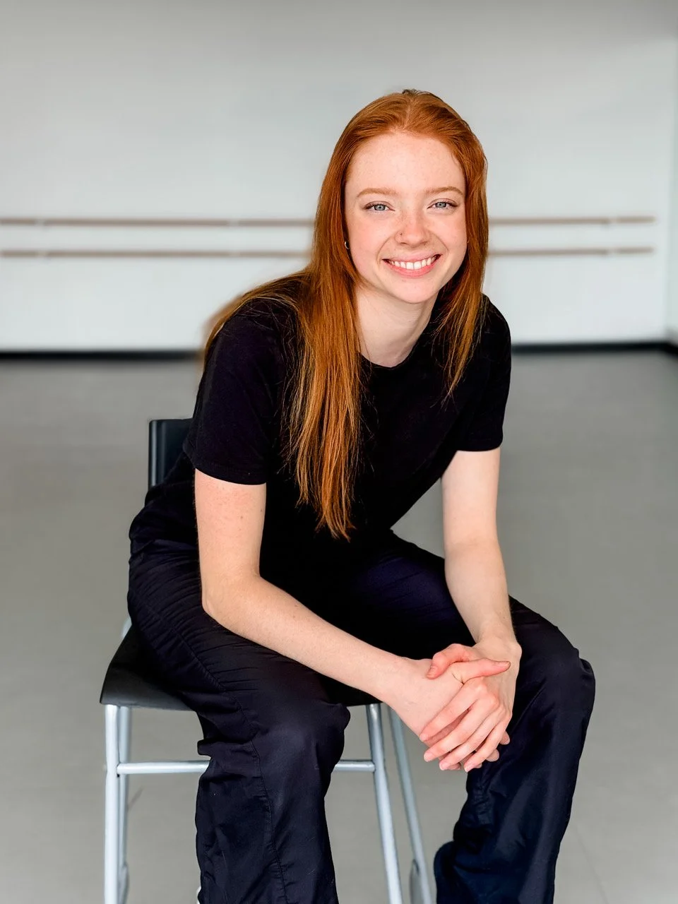 Young woman with long red hair wearing black t-shirt and pants, sitting on a chair in a room with a plain background