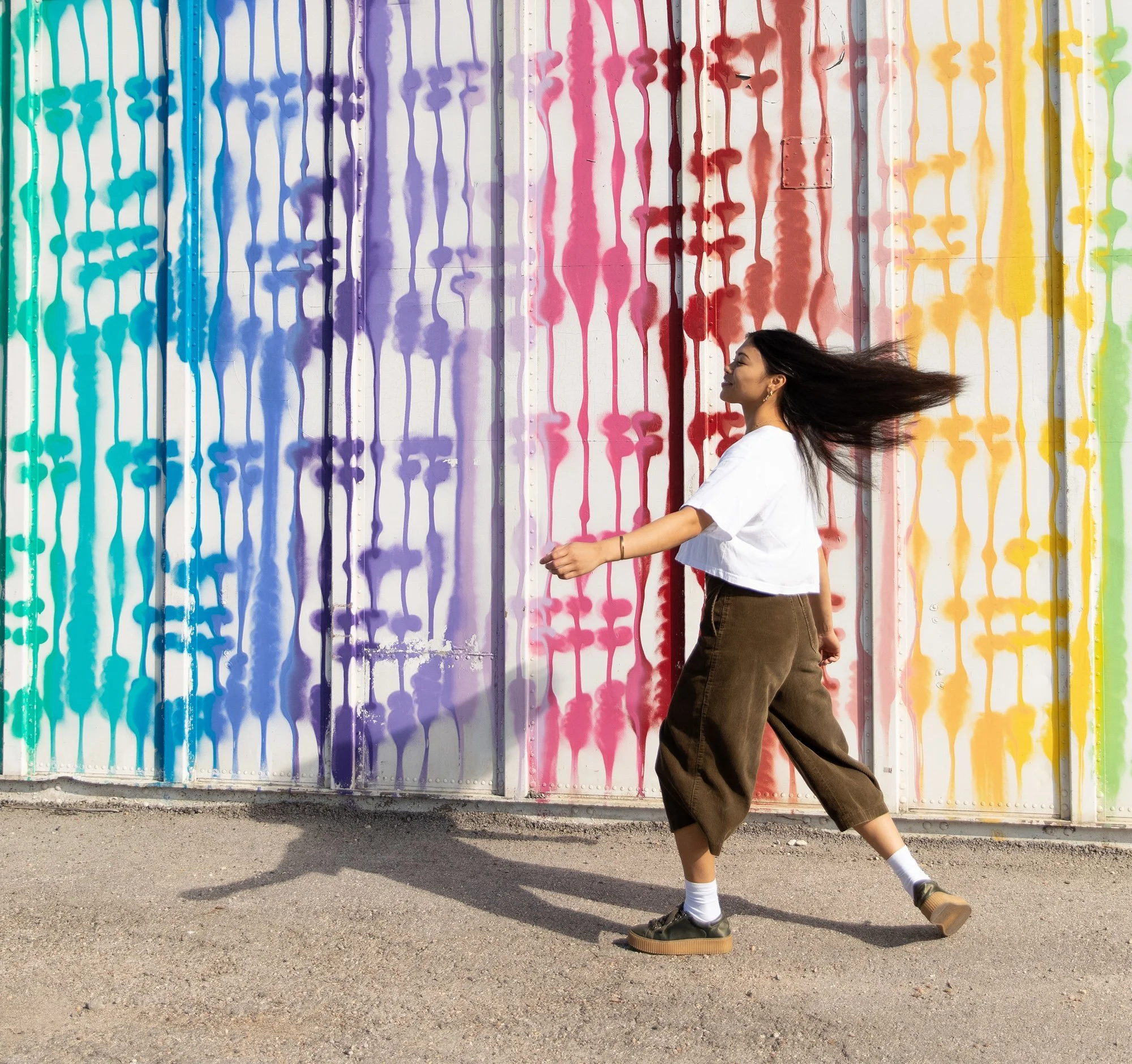 A woman with long dark hair, wearing a white t-shirt and brown wide-leg pants, walking past a colorful graffiti wall with rainbow colors in the background.