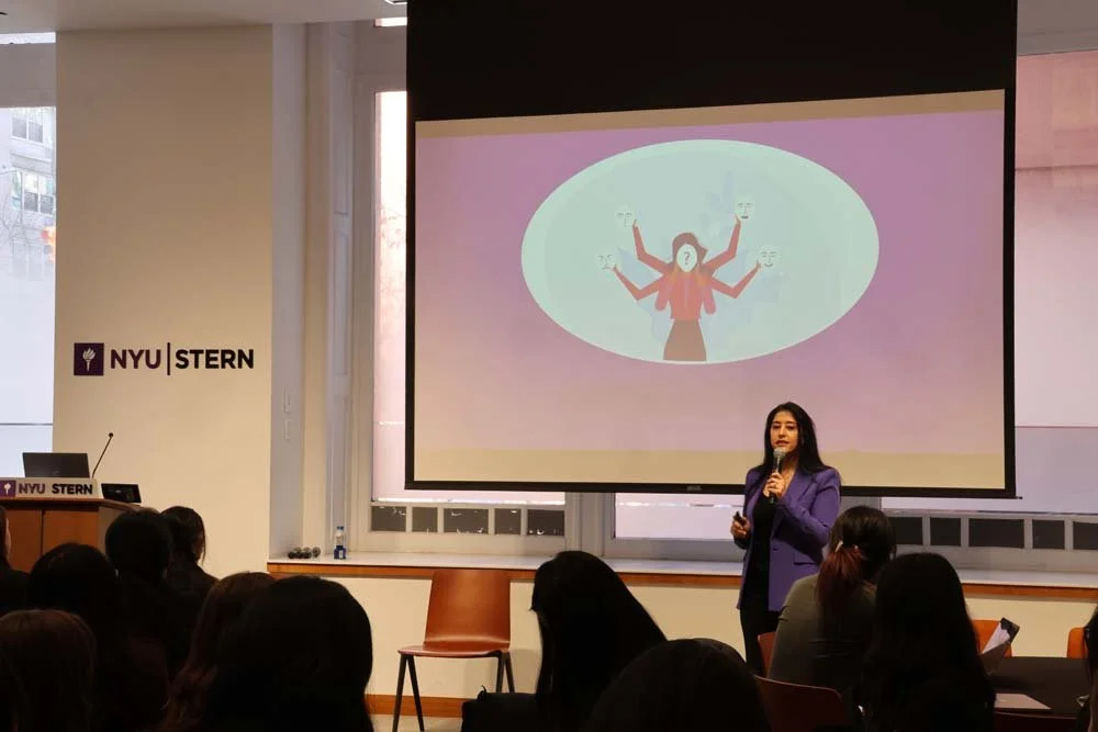 A woman in a professional purple blazer giving a presentation with a microphone in a classroom at NYU Stern School of Business. Behind her is a large screen displaying a cartoon of a person with multiple arms holding different objects.