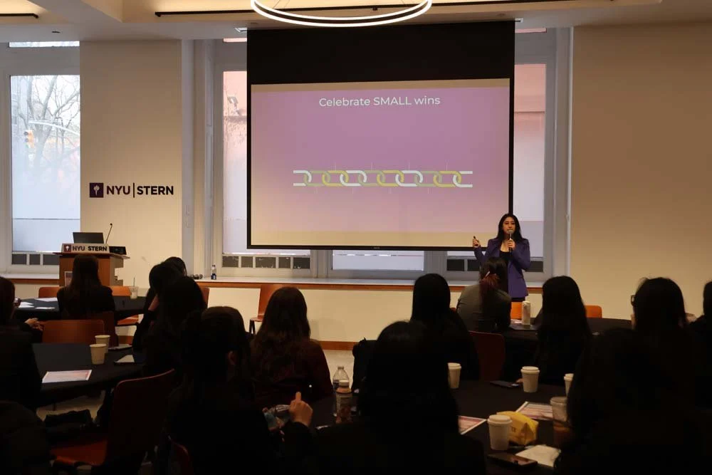 A woman giving a presentation at NYU Stern School of Business, with a slide that reads 'Celebrate SMALL wins', in a room with an audience and large windows.