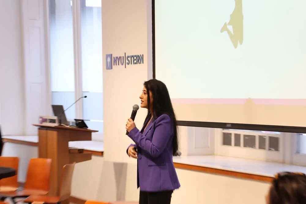 A woman in a purple blazer holding a microphone and speaking in front of a presentation screen with NYU Stern logo in a conference room.