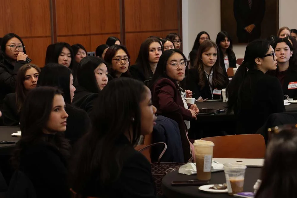 Group of diverse young women attending a conference or seminar in a large room, seated at tables with beverages and notepads.