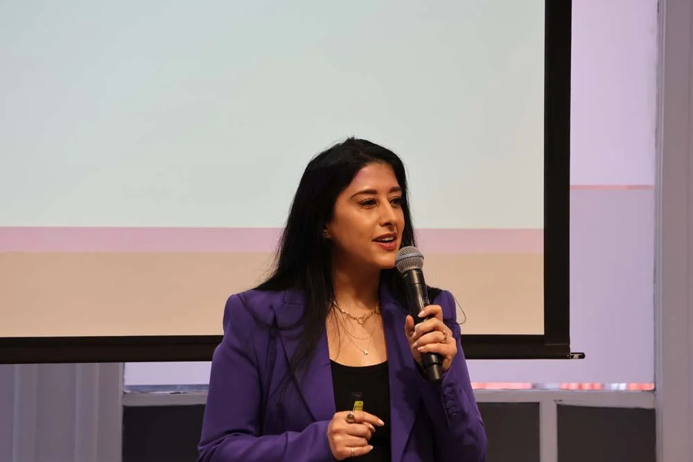 A woman in a purple blazer speaking into a microphone in front of a projection screen.