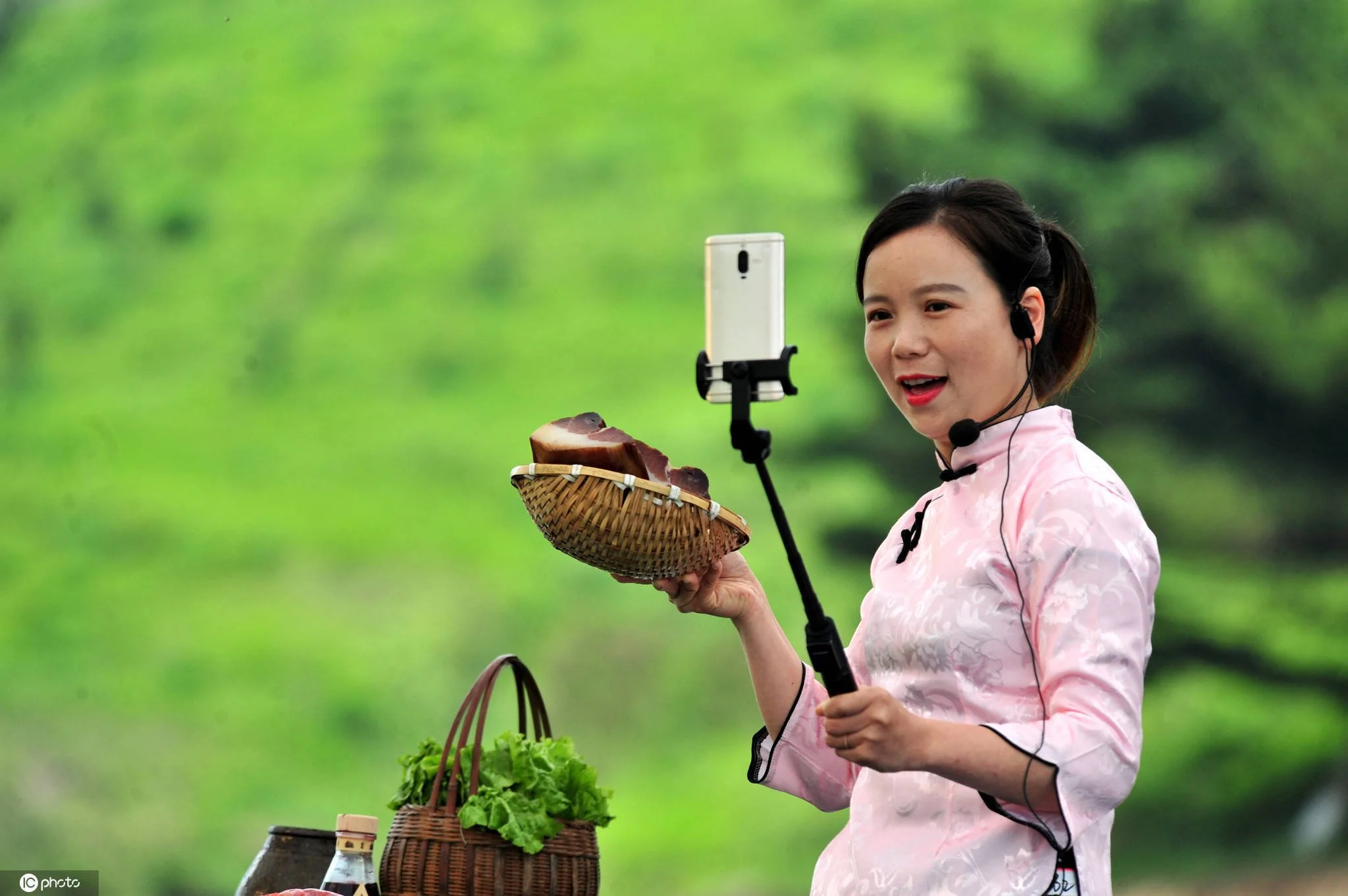 A woman with a pink traditional dress takes a selfie with a smartphone mounted on a selfie stick outdoors, holding a basket of herbs or vegetables, with a green, blurred background.