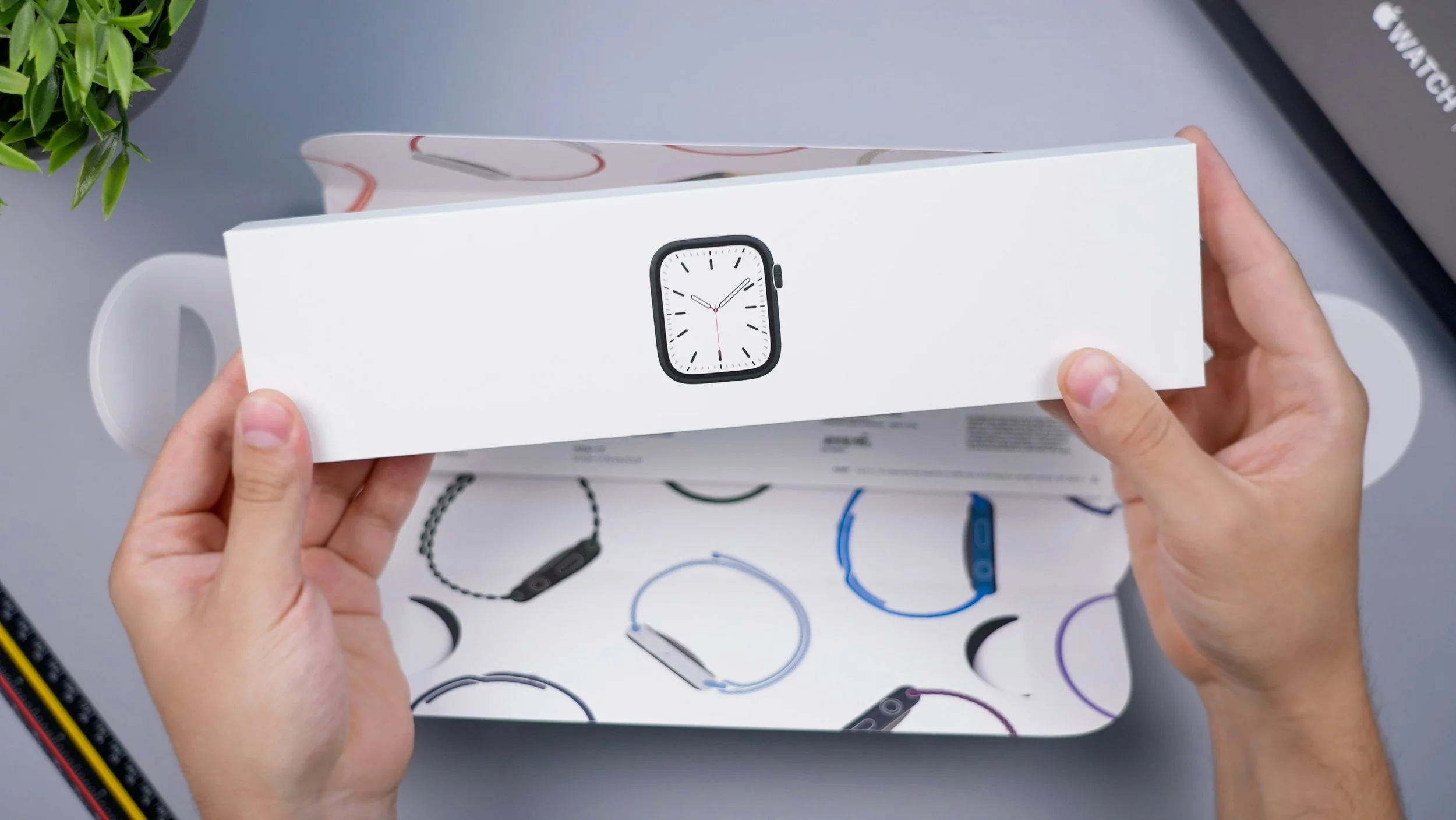 Person holding a white box with an Apple Watch icon on it, with a partially opened box underneath showing illustrations of various Apple Watch bands.