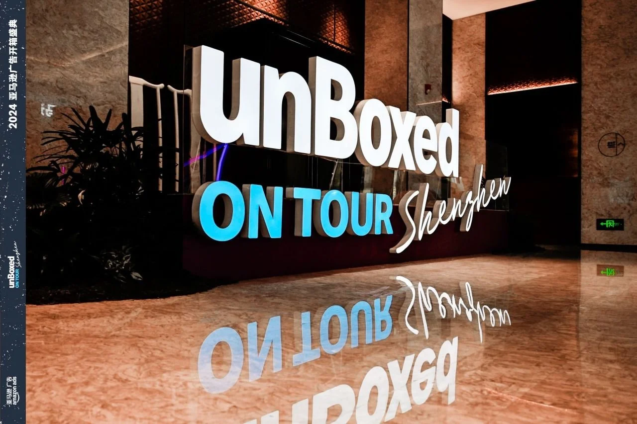 Large illuminated sign inside a building reads 'UnBoxed ON TOUR Shaman' with a reflection on the polished floor.