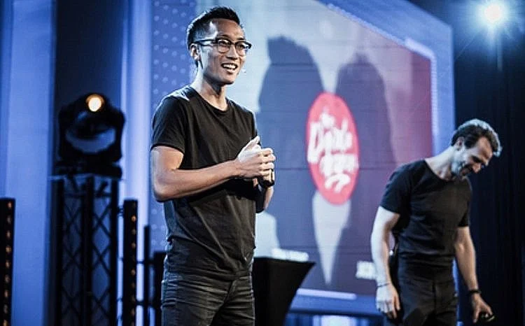 Two men on stage, one smiling and holding a microphone, with stage lights and a large screen in the background.