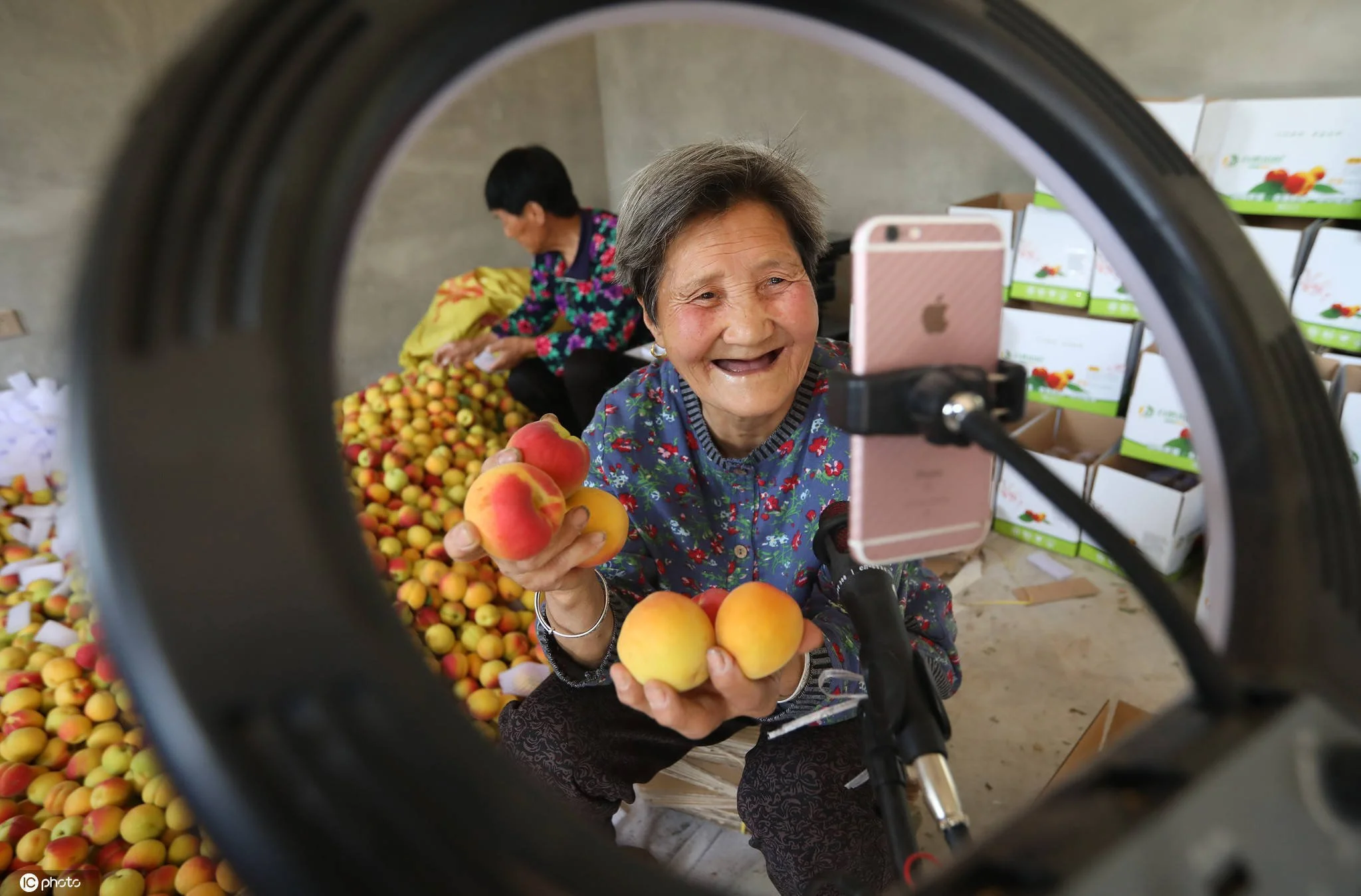 An elderly woman is taking a selfie with her smartphone, holding peaches, at a fruit market with a woman in the background sorting through red and yellow apricots